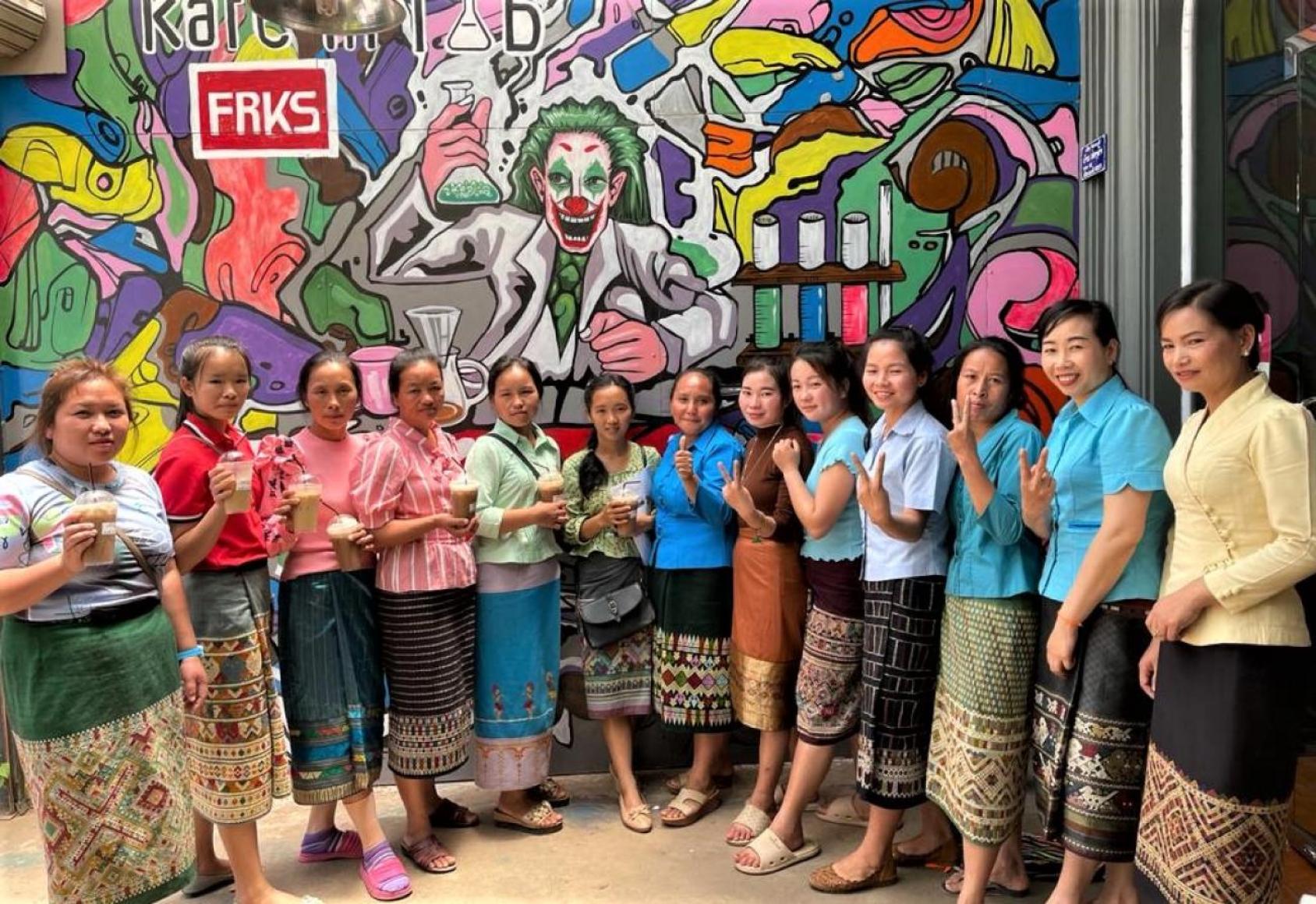 Plusieurs femmes vêtues de tenues laotiennes se tiennent debout en demi cercle, devant un mur recouvert d'une fresque aux couleurs vives.