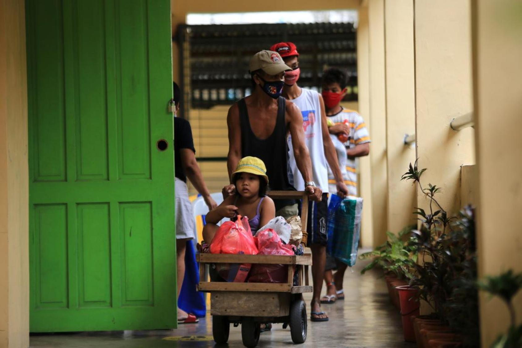 La photo montre plusieurs personnes dont un enfant portant des masques de protection respiratoire à l'intérieur d'un marché et transportant des sacs de produits alimentaires. 