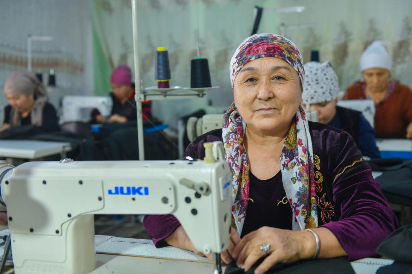 In a room full of women working on their sewing machines a woman looks directly at the camera. 