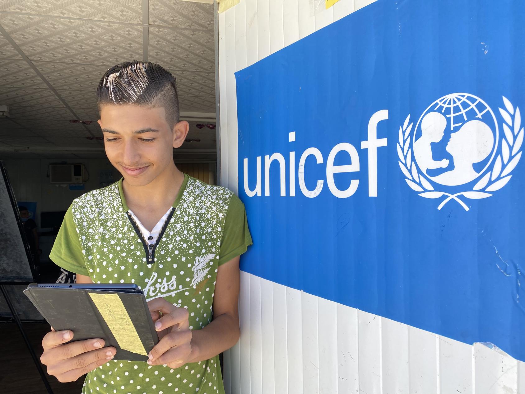 A boy smiles while he looks down at his tablet near a blue UNICEF sign. 