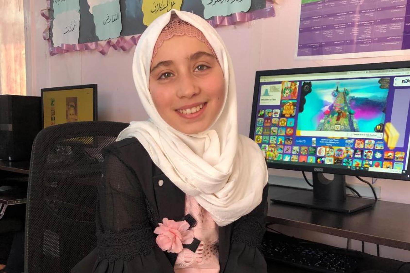 A smiling girl, with a white hijab, smiles at the camera near a computer displaying a game. 