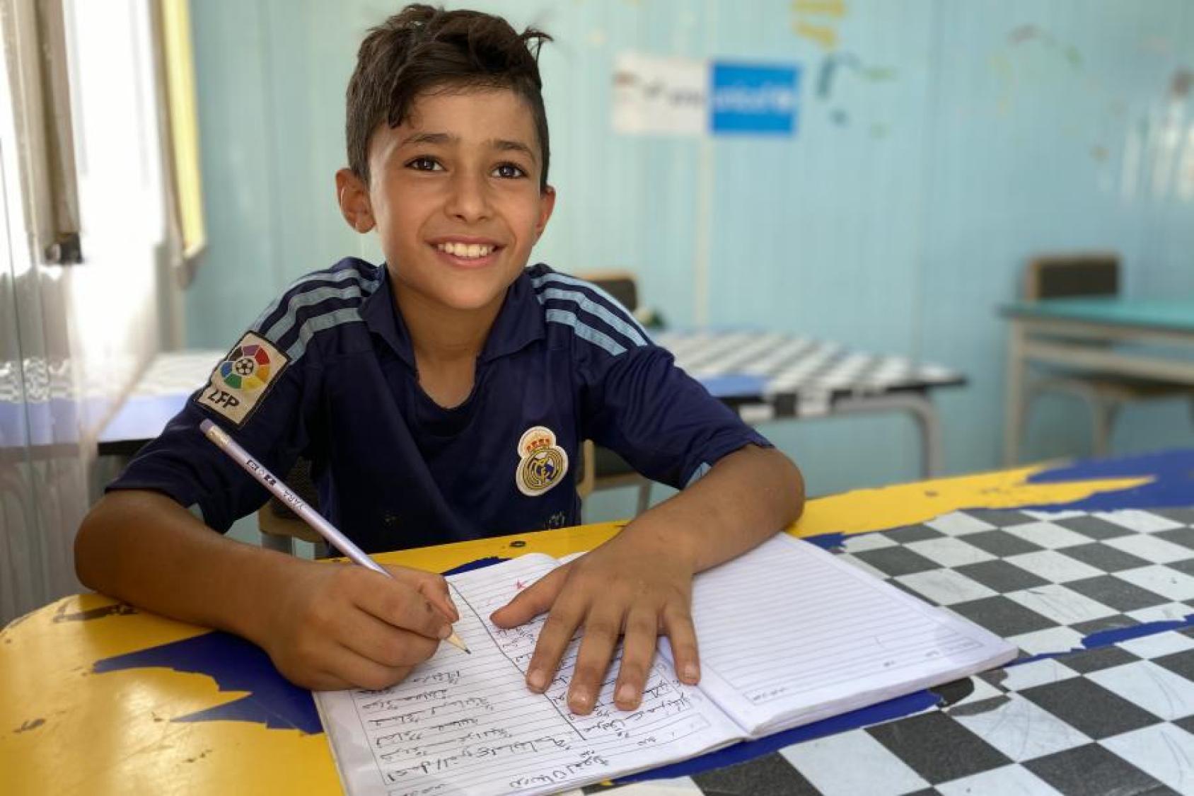 A young boy, wearing a blue jersey, writes in a notebook with a pencil. 