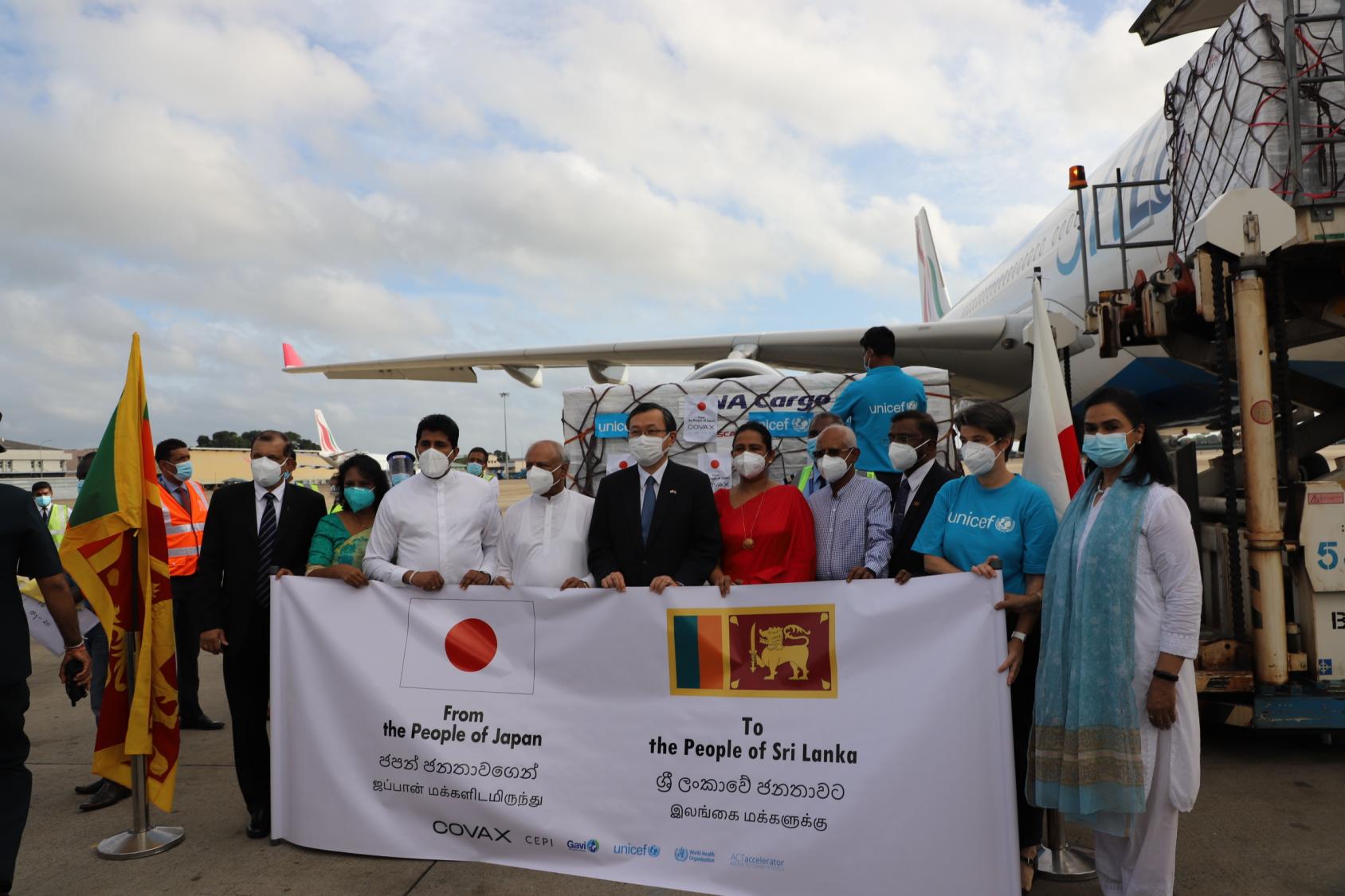 A group of people hold a banner with the Japanese and Sri Lankan flags in front of an airplane and a delivery of COVAX vaccines. 