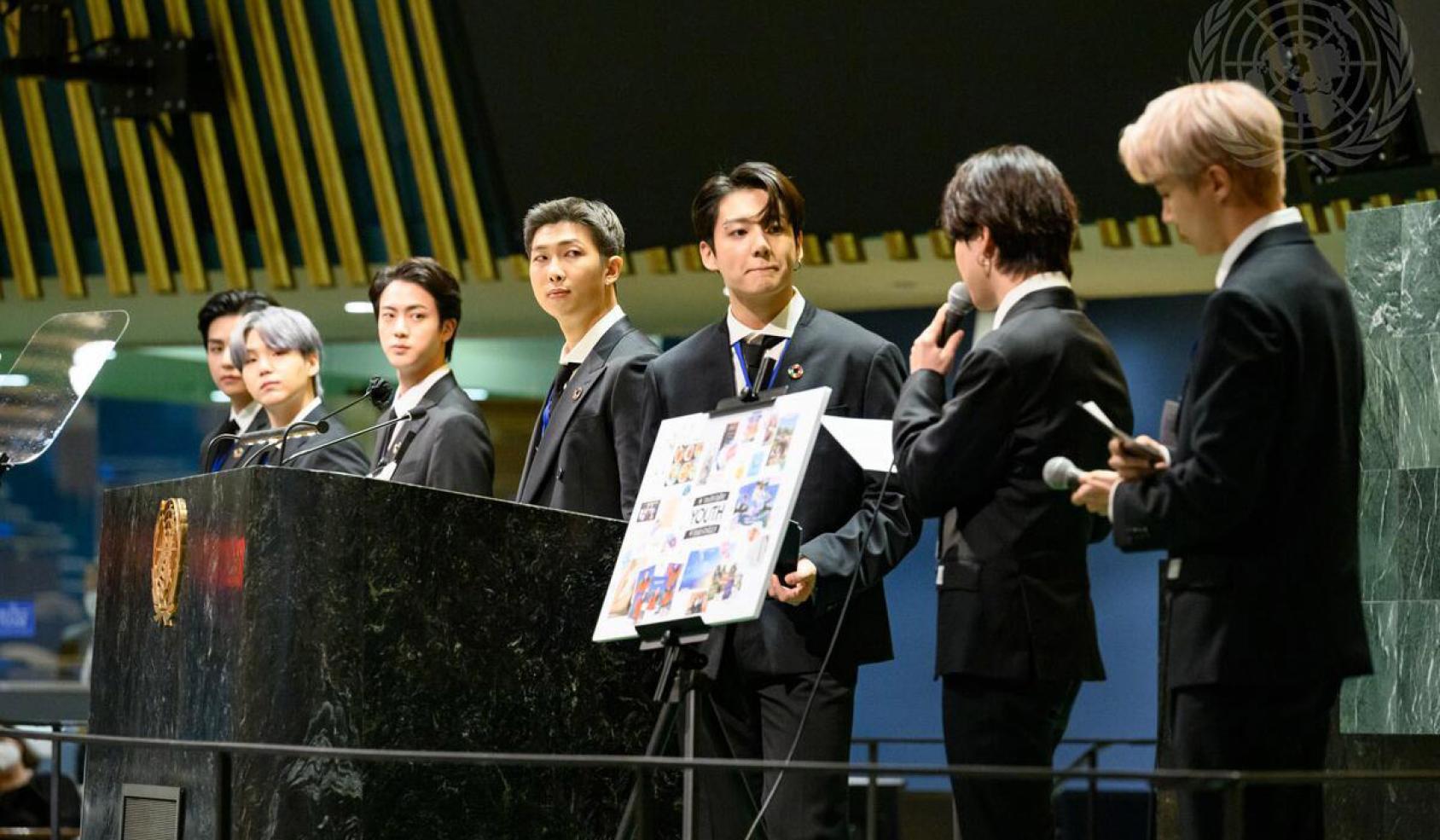 Members of the band, BTS, stand near a sign on the stage of the General Assembly Hall as they perform. 