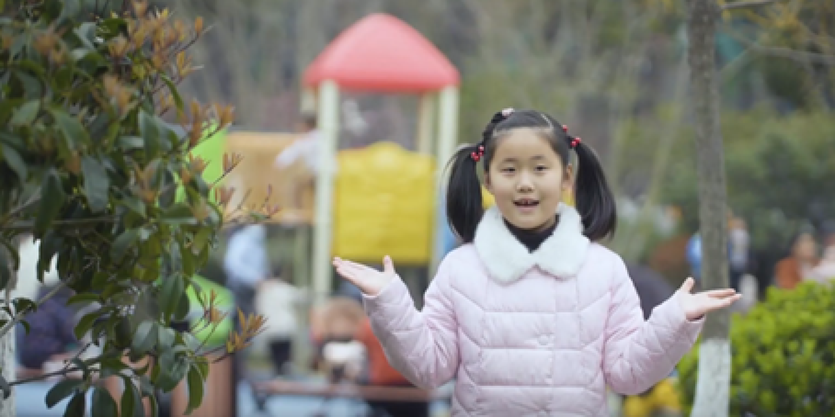 A girl in a pink jacket stands near a playground. 