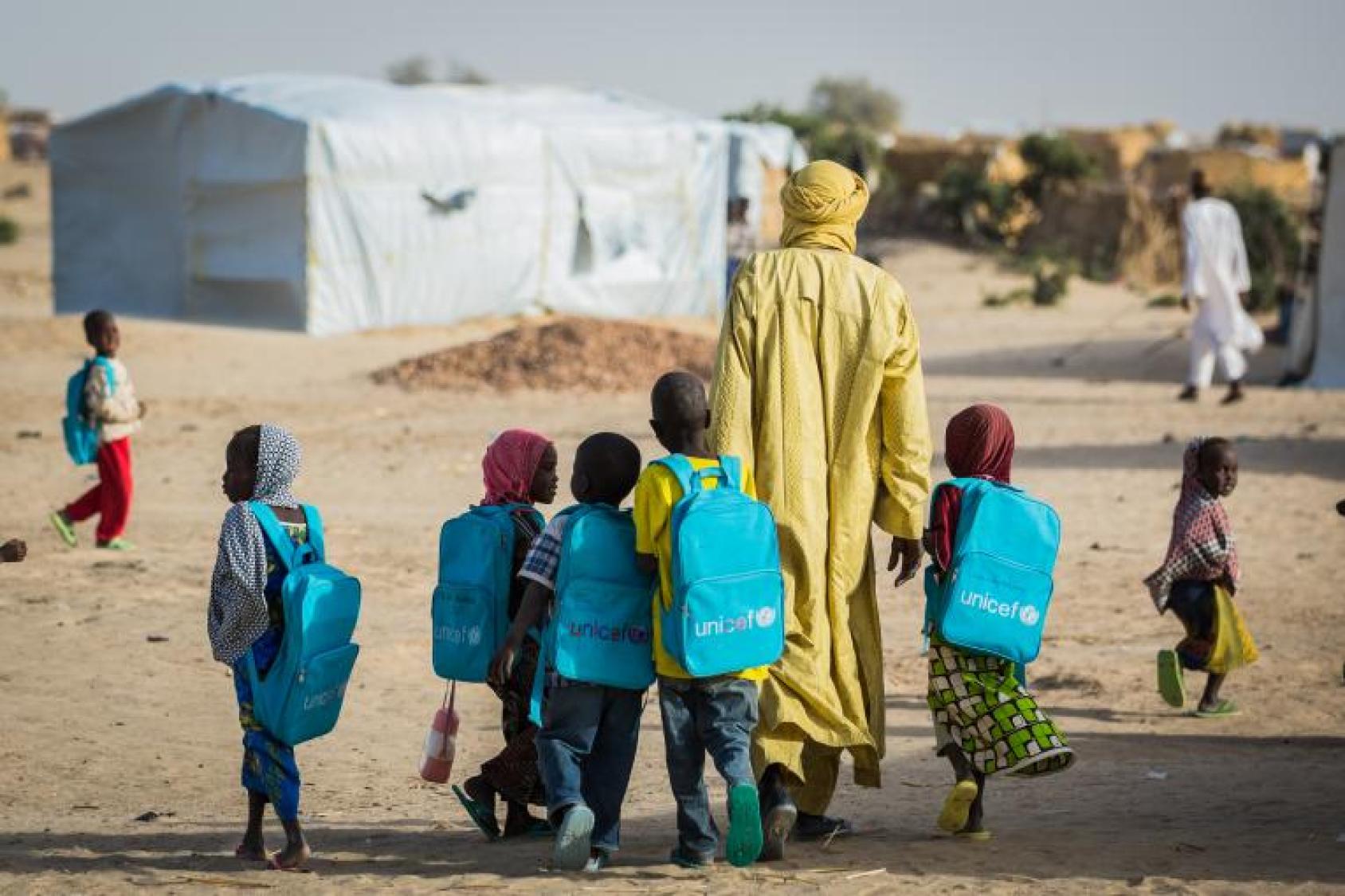 Un grupo de niños y niñas llevan mochilas azules de UNICEF mientras caminan con un adulto cerca de las tiendas de campaña.