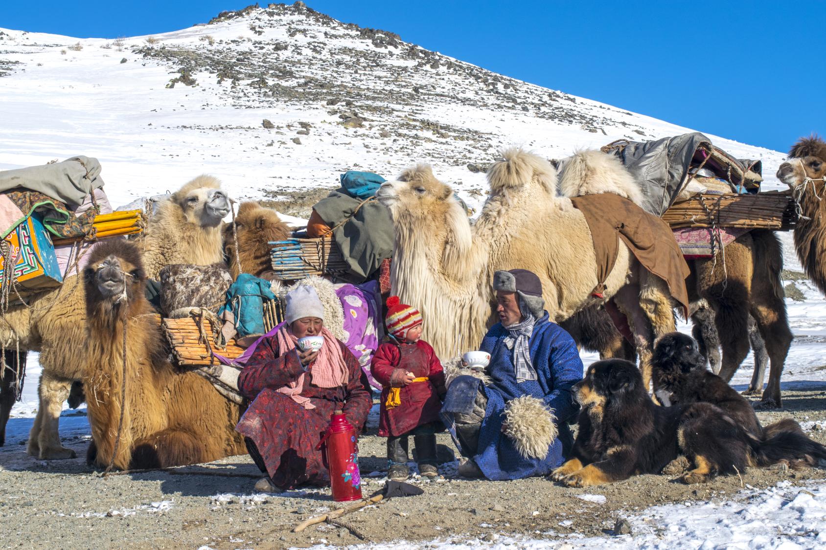 Une famille nomade de Mongolie composée d'un homme, d'une femme et d'un petit enfant emmitouflés dans des vêtements épais se repose en buvant du thé, entourée de deux chiens noirs et de plusieurs chapeaux. En arrière-plan s'élève une colline enneigée.