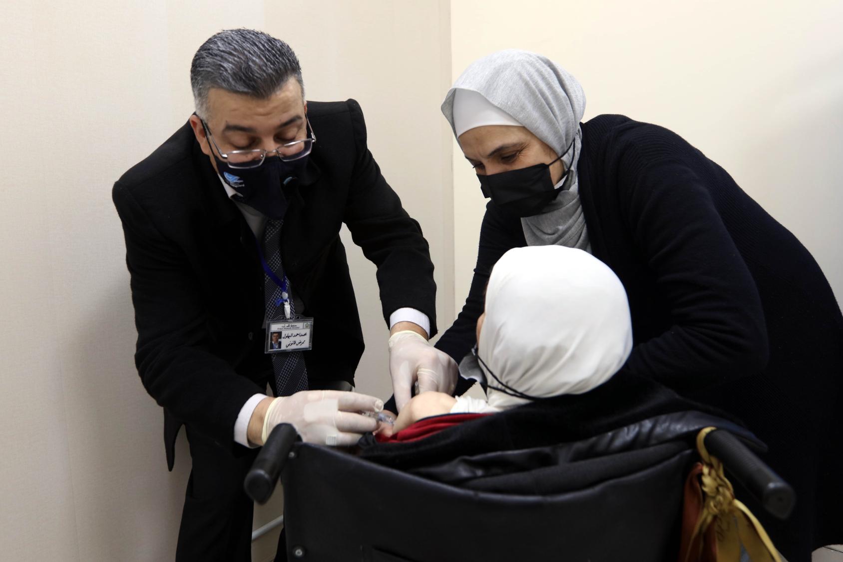 Une femme en fauteuil roulant se fait administrer un vaccin anti-COVID-19 par un homme vêtu d'un costume et portant un masque de protection respiratoire noir et des gants de protection. Une autre femme, voilée et portant elle aussi un masque de protection respiratoire noir aide l'homme à administrer le vaccin à la patiente.