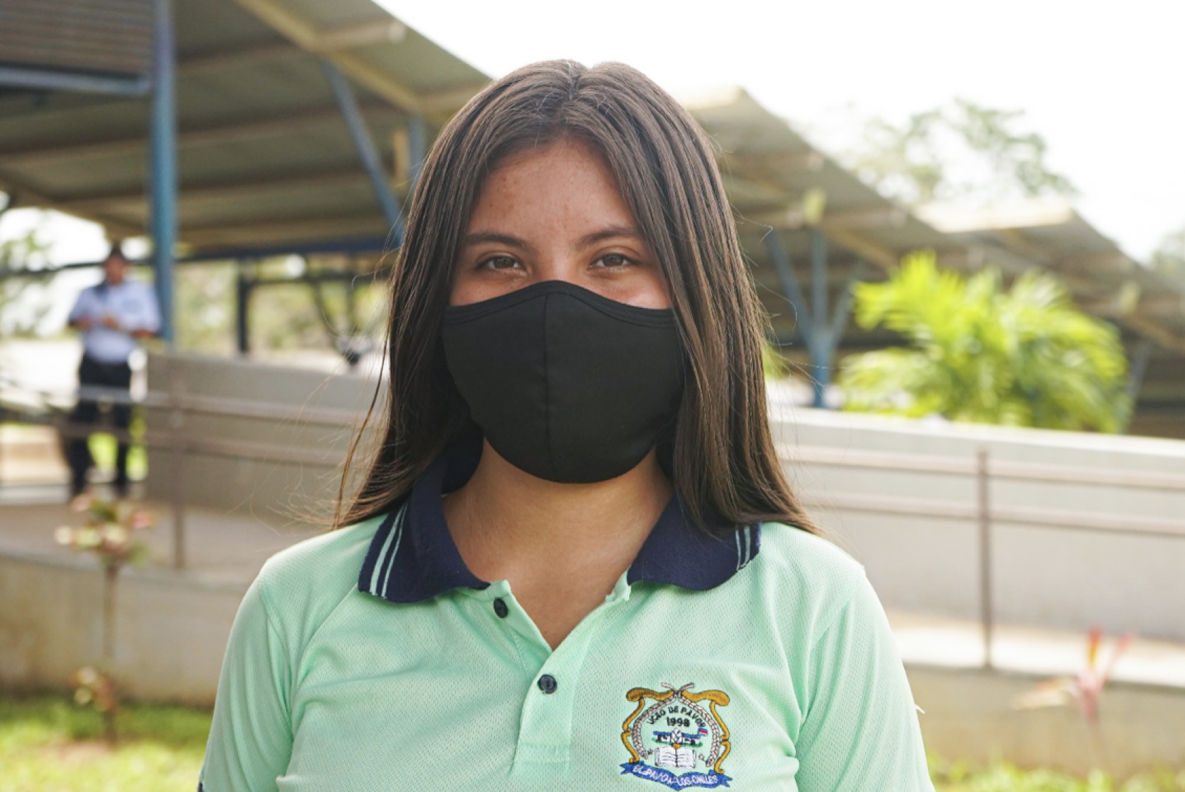 A girl in a school uniform and black face mask looks directly at the camera at her school. 