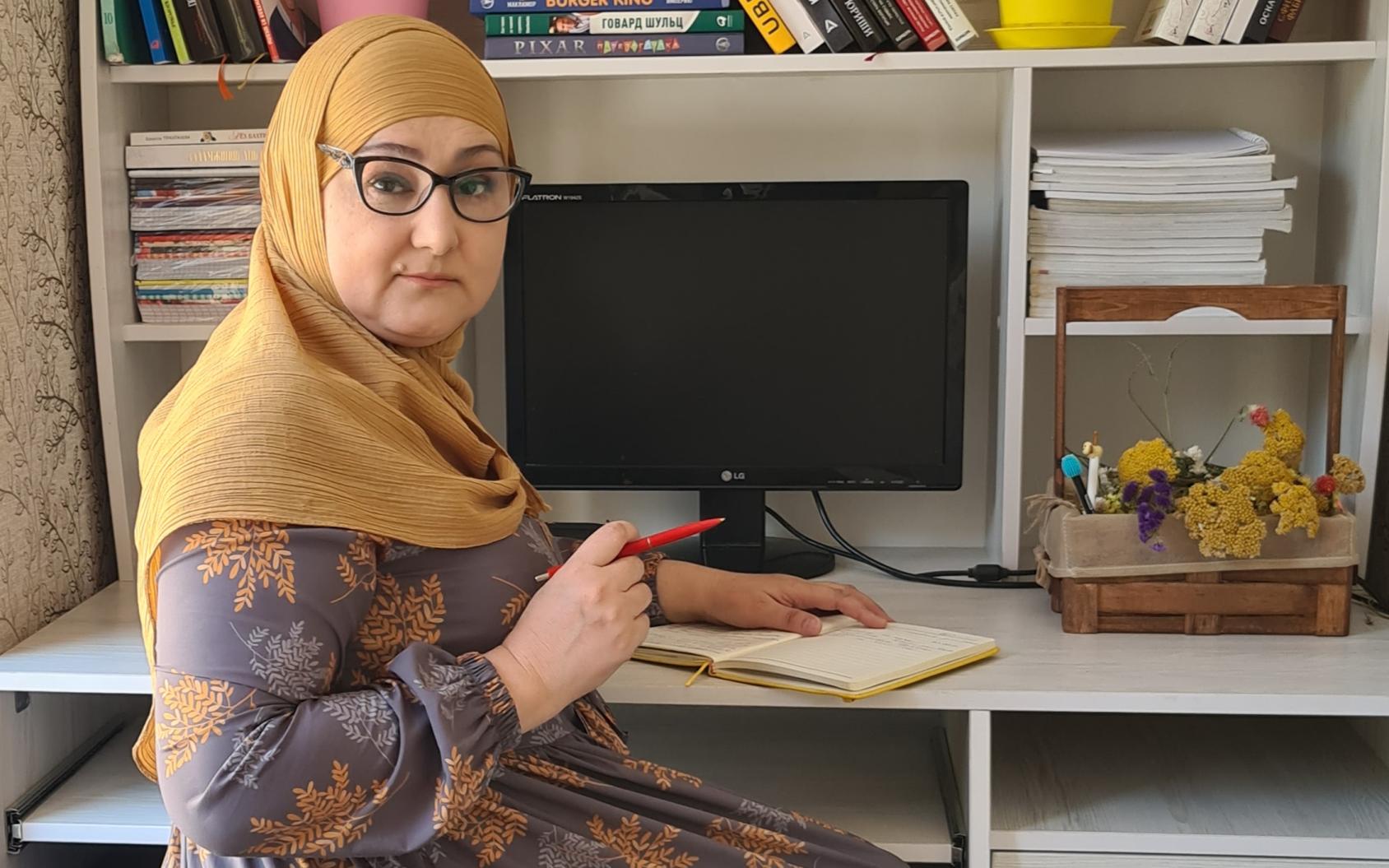 Una mujer, en la oficina de su casa con un ordenador, libros y papeles, escribe en un diario.