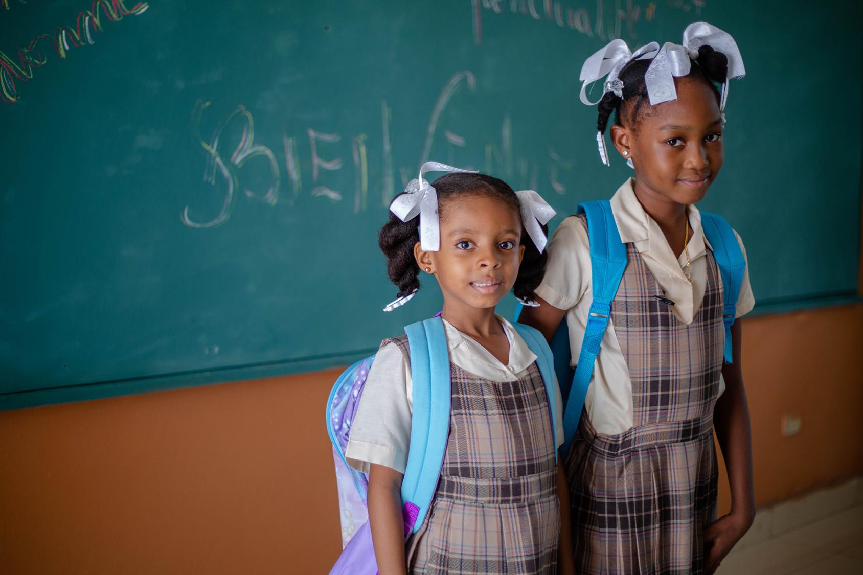 Dos niñas con mochila y uniforme escolar se sitúan frente a una pizarra.