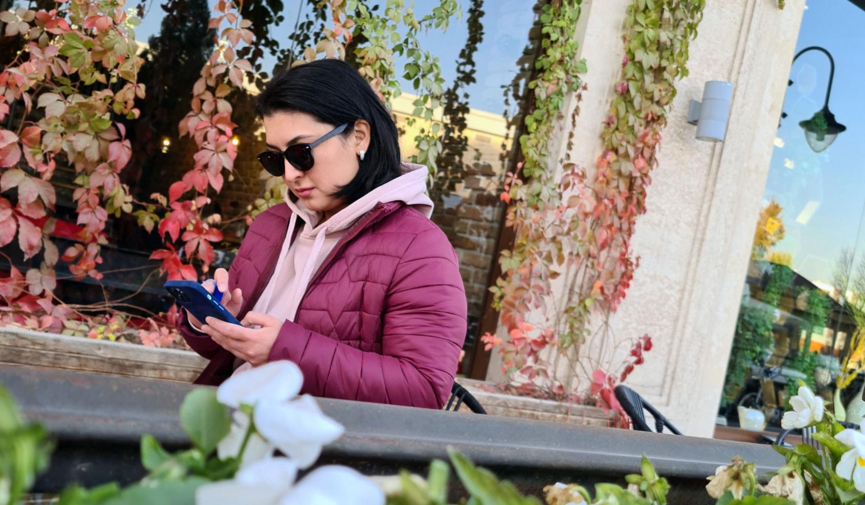 Aqida Mokhirova stands reading messages on her mobile phone as she stands outside a local business window covered with blooming flowers and pink vines.