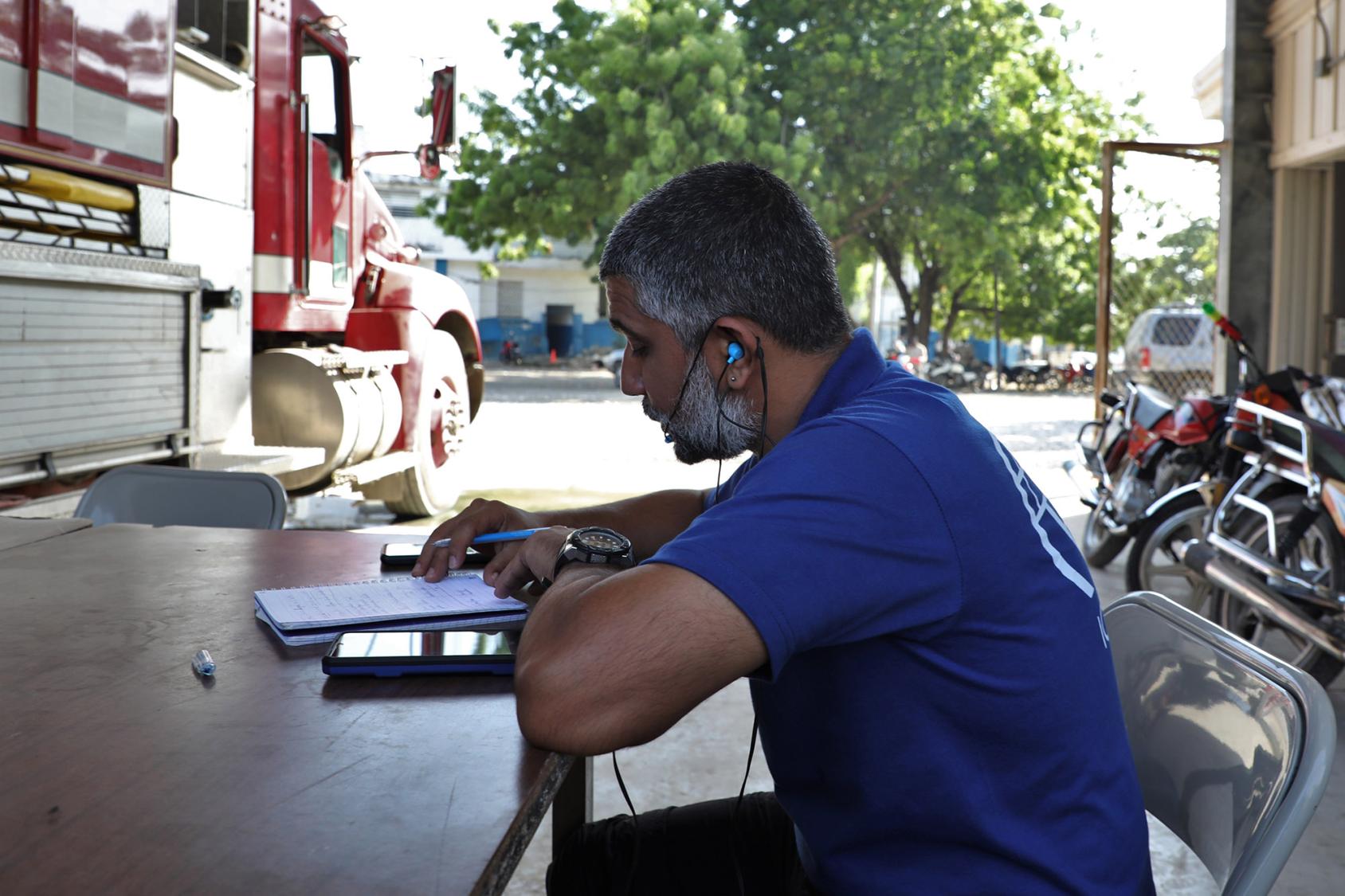 A man in a blue shirt looks down at a mobile phone and paperwork inside a fire station. 