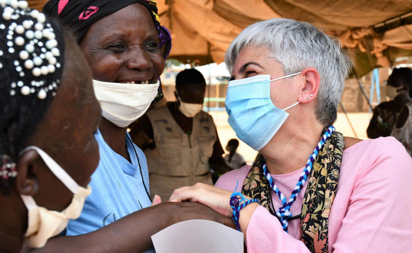Resident and Humanitarian Coordinator in Burkina Faso Barbara Manzi (right) smiles and holds the hand of a smiling woman, wearing a mask, while another one faces them on the left. 