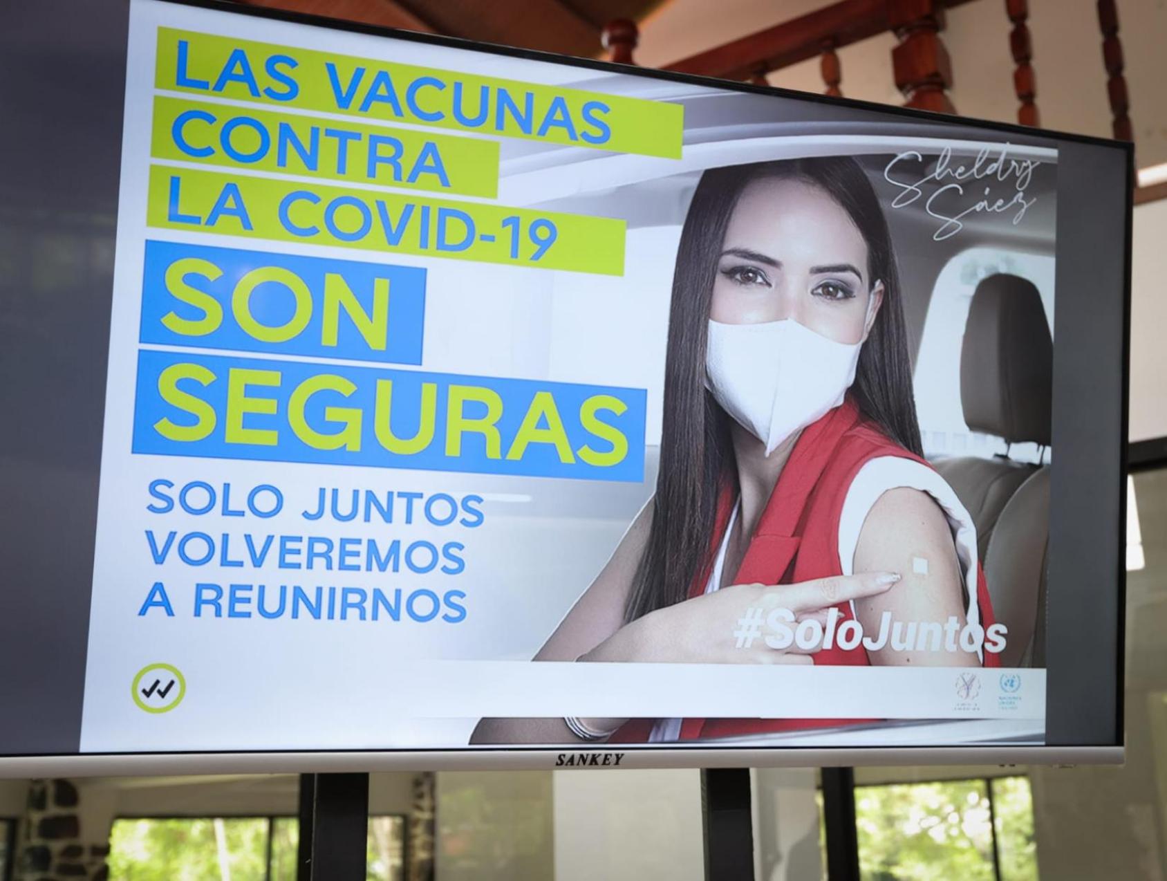 一个戴着面具的妇女指着她手臂上的创可贴，旁边是西班牙语的 "新冠疫苗是安全的 "字样的标志。
