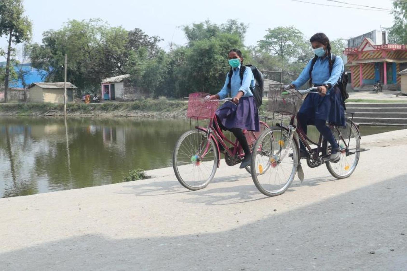 Two young girls in school uniforms ride bikes down a dirt path near a body of water. 