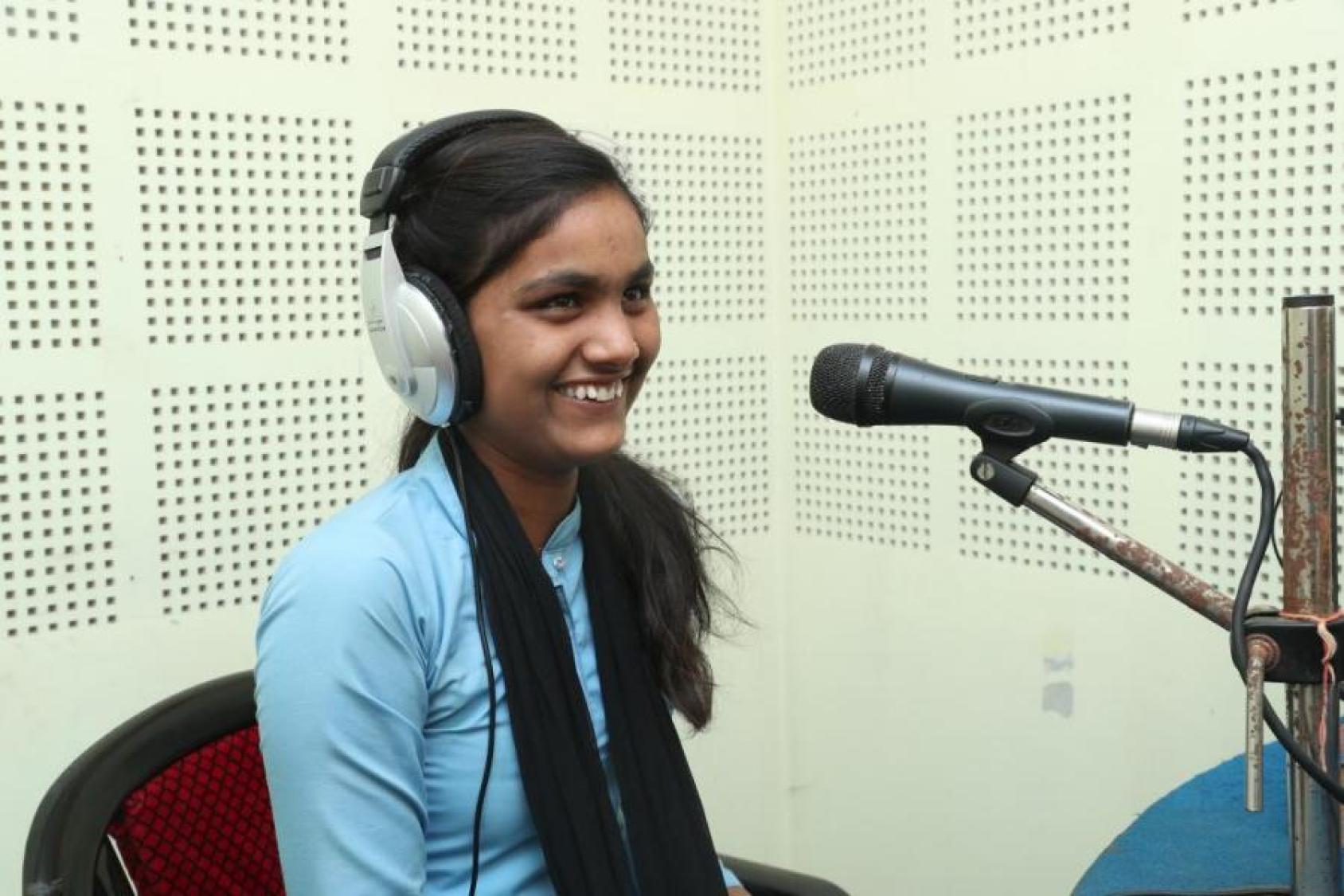 A girl in a studio has headphones on and smiles at the microphone near her mouth.