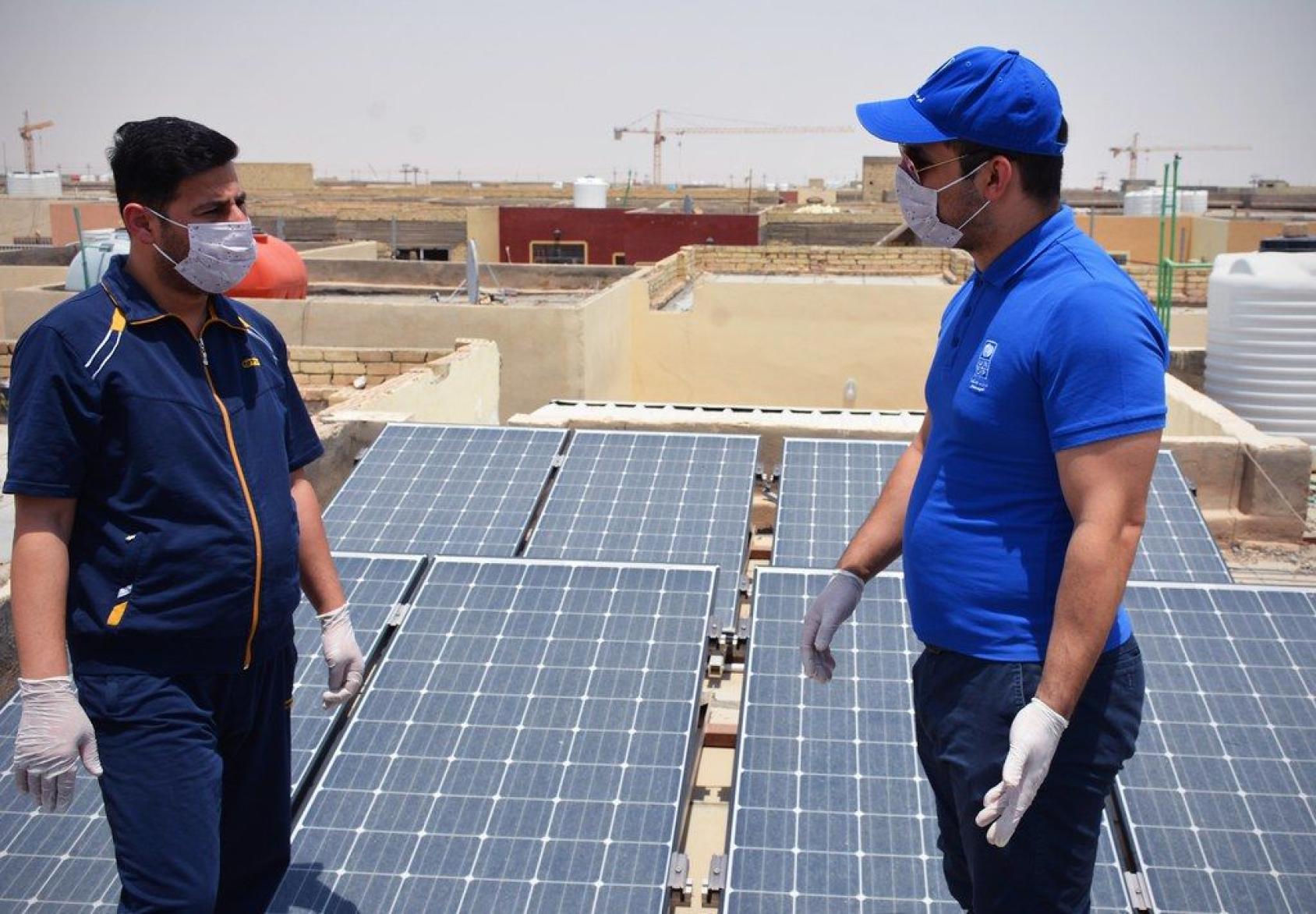 Two men in face masks and gloves stand near solar panels on a sunny day.