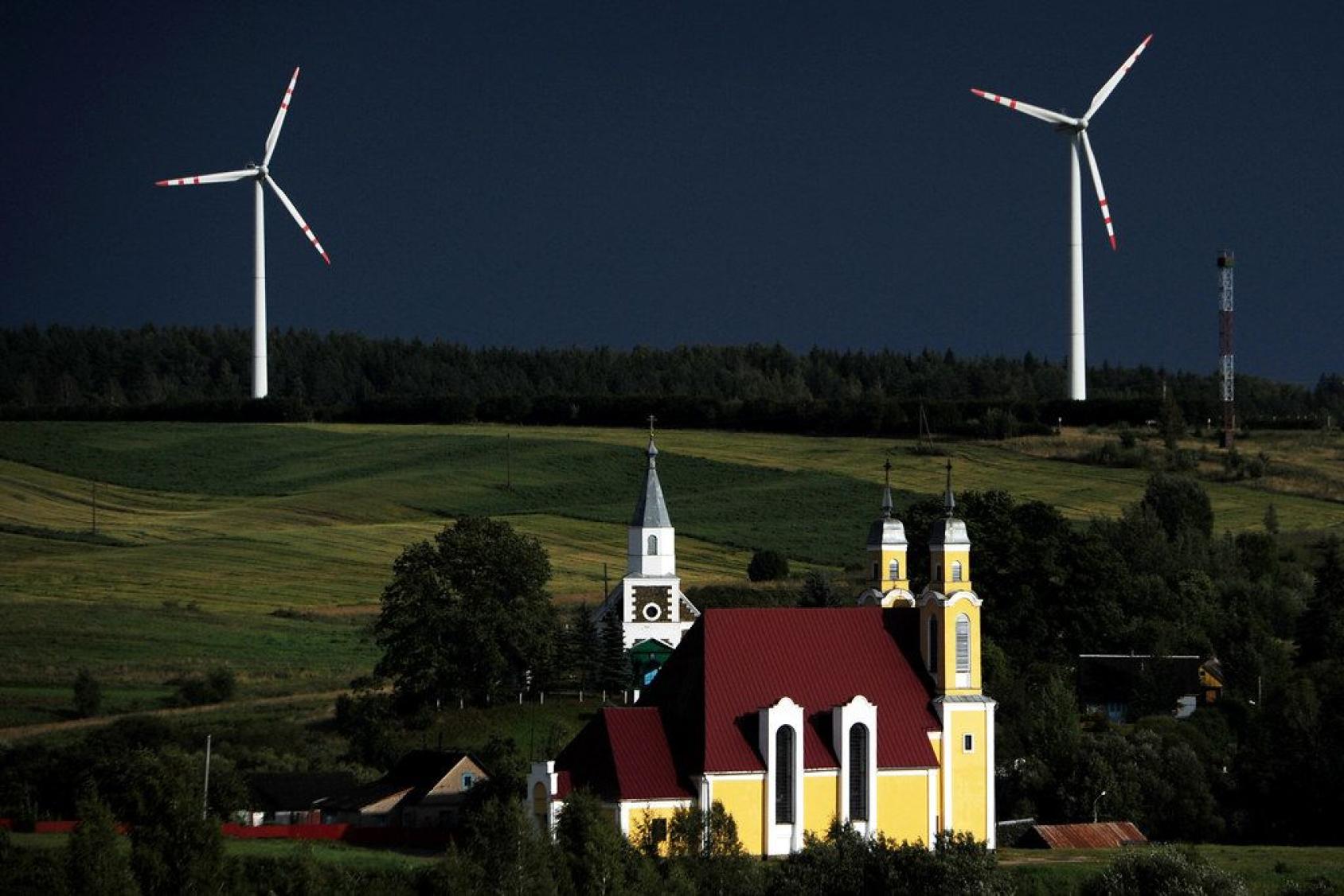 A landscape image of wind turbines behind a yellow church at sunset. 