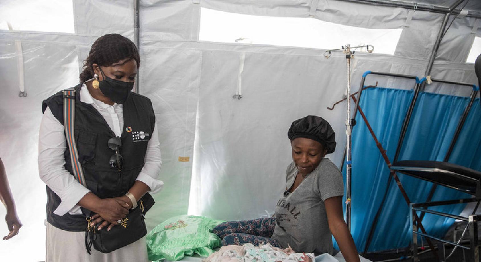 UNFPA Deputy Executive Director, Diene Keita, stands next to a mother who is sitting on a bed next to her new baby.