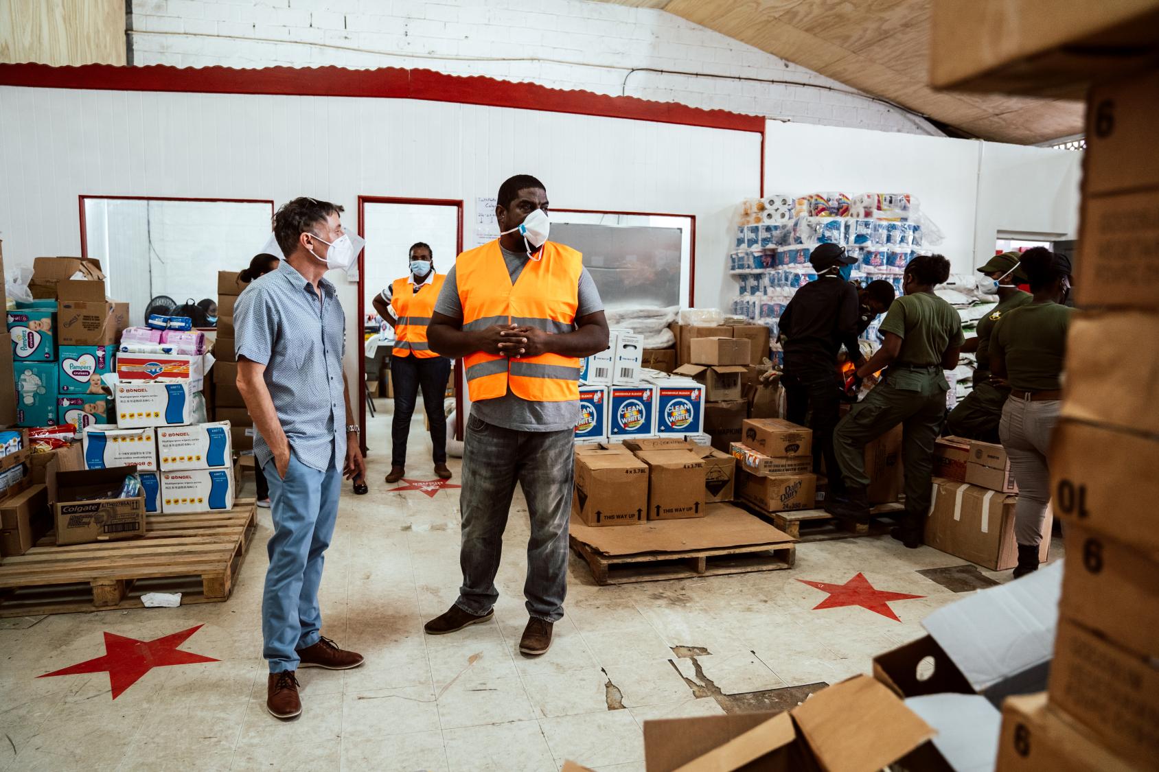 The resident coordinator stands with another man in a warehouse and several people work behind them. 