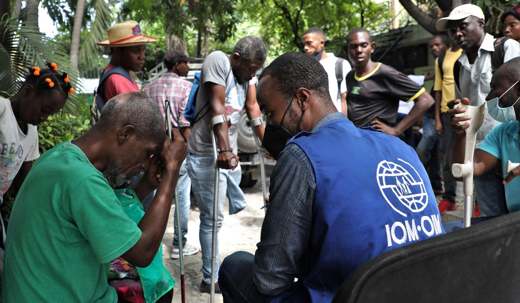 An IOM staff member sits beside a man holding white cane, while other men of various abilities stand in the background. 
