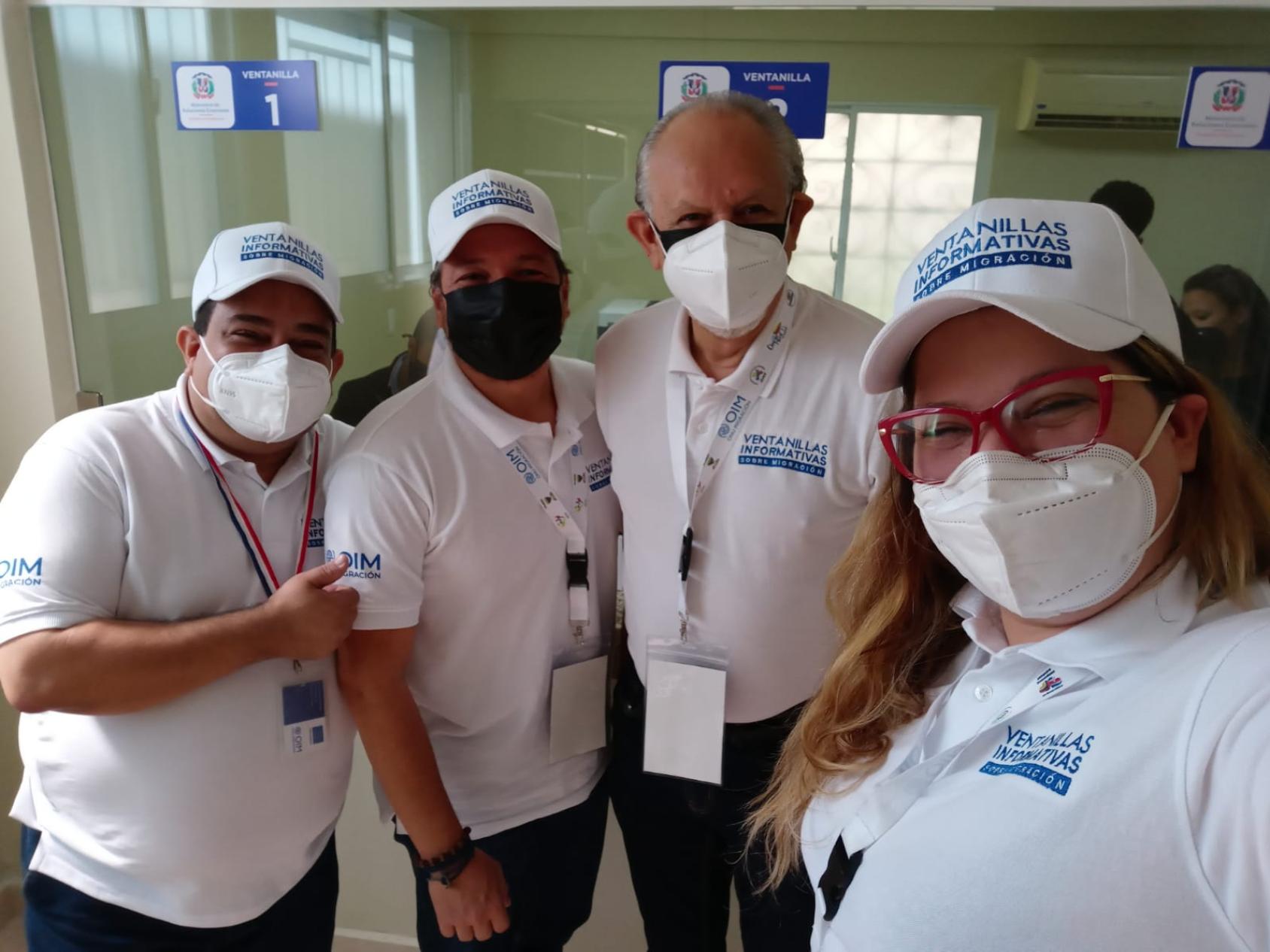 Three men and a woman, all wearing white IOM embroidered t-shirts and face mask, smile and pose for a selfie.