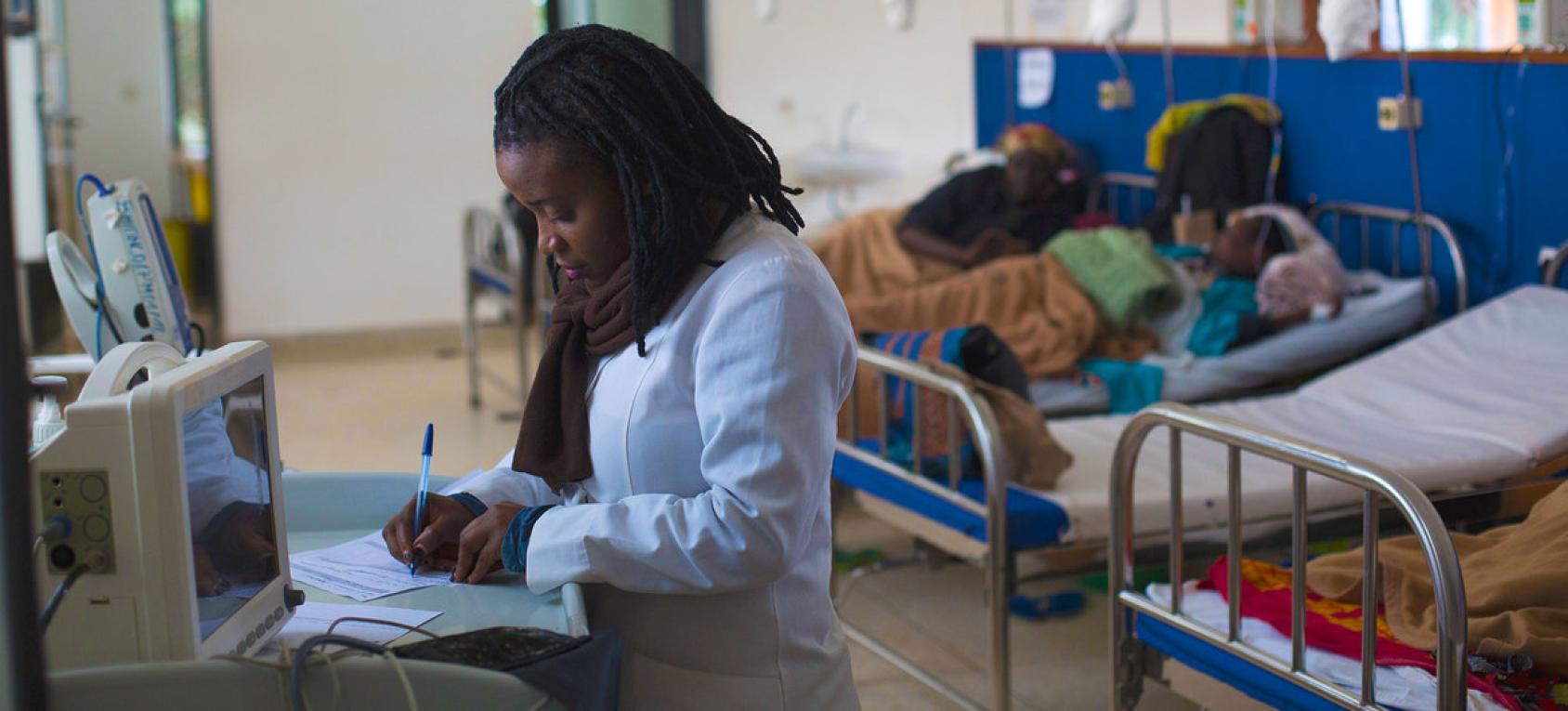 A nurse writes on a form as she stands by a computer desk with patients in beds in the background.