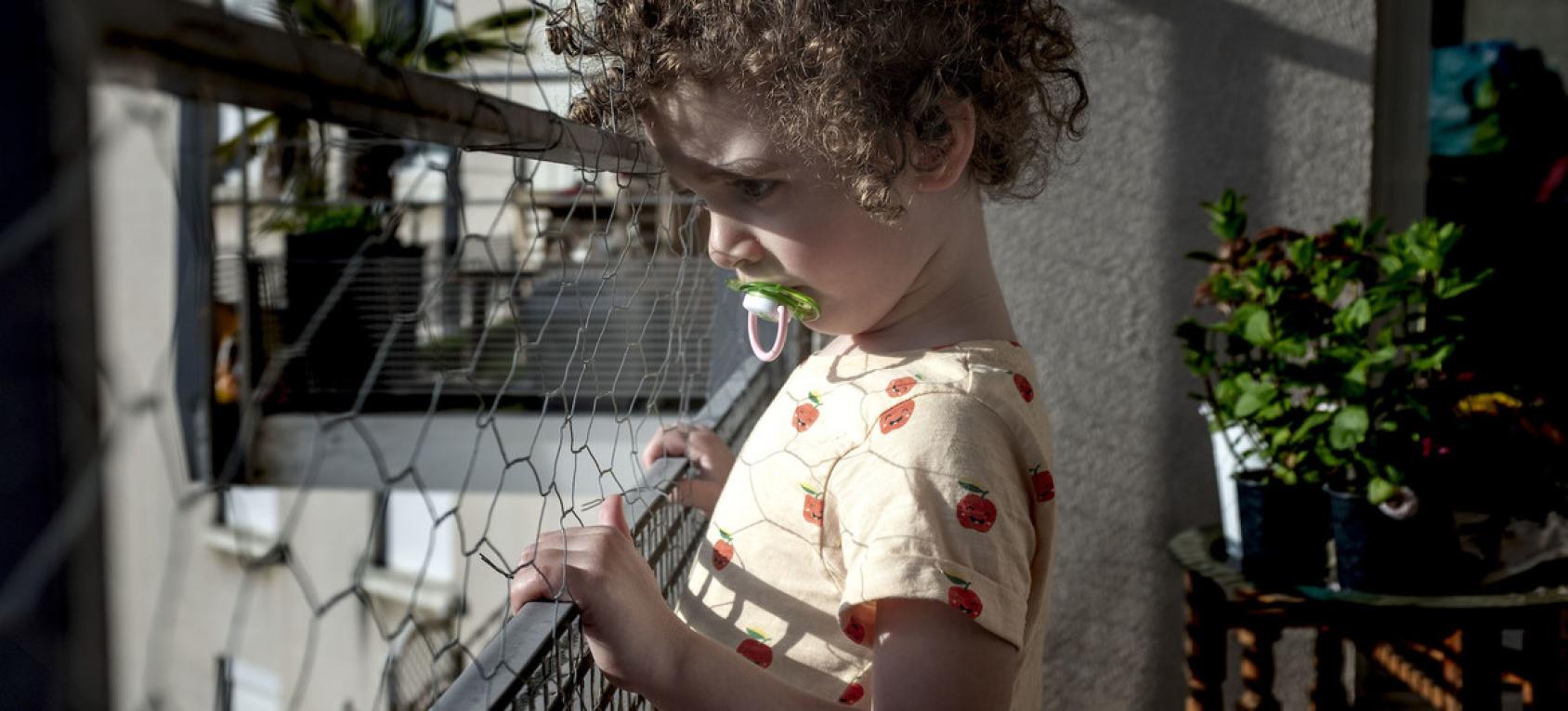 A curly haired little girl with a pacifier looks outside a gate on the porch of her apartment building.
