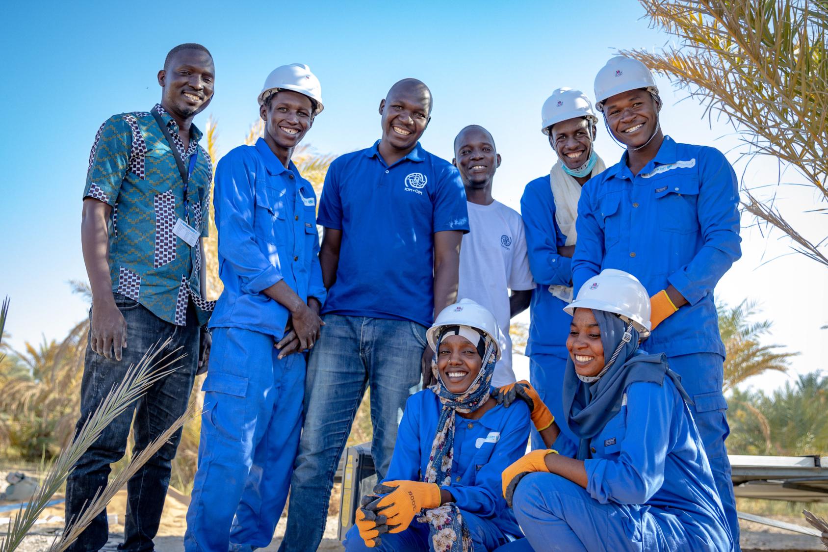 Группа молодых людей в синих рабочих комбинезонах улыбается в камеру.
