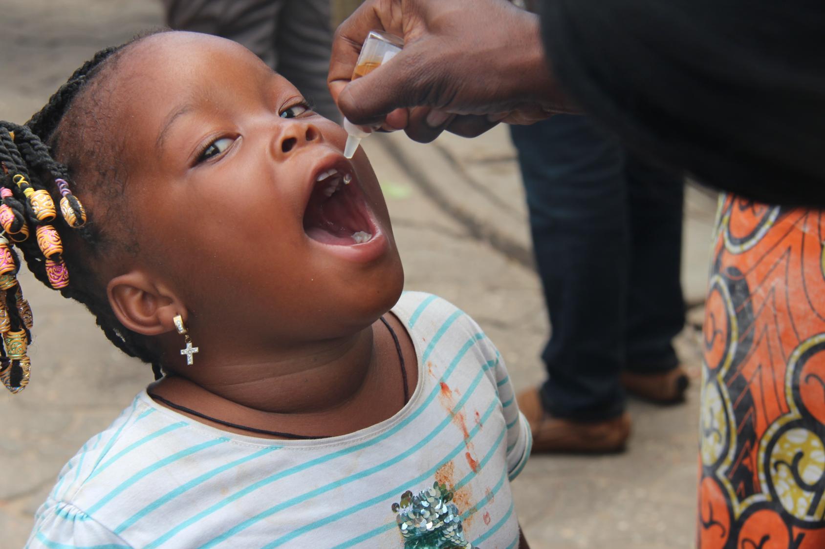 Une petite fille béninoise coiffée avec des tresses ouvre la bouche tandis qu'un adulte lui administre le vaccin de la polio sous forme de gouttes.