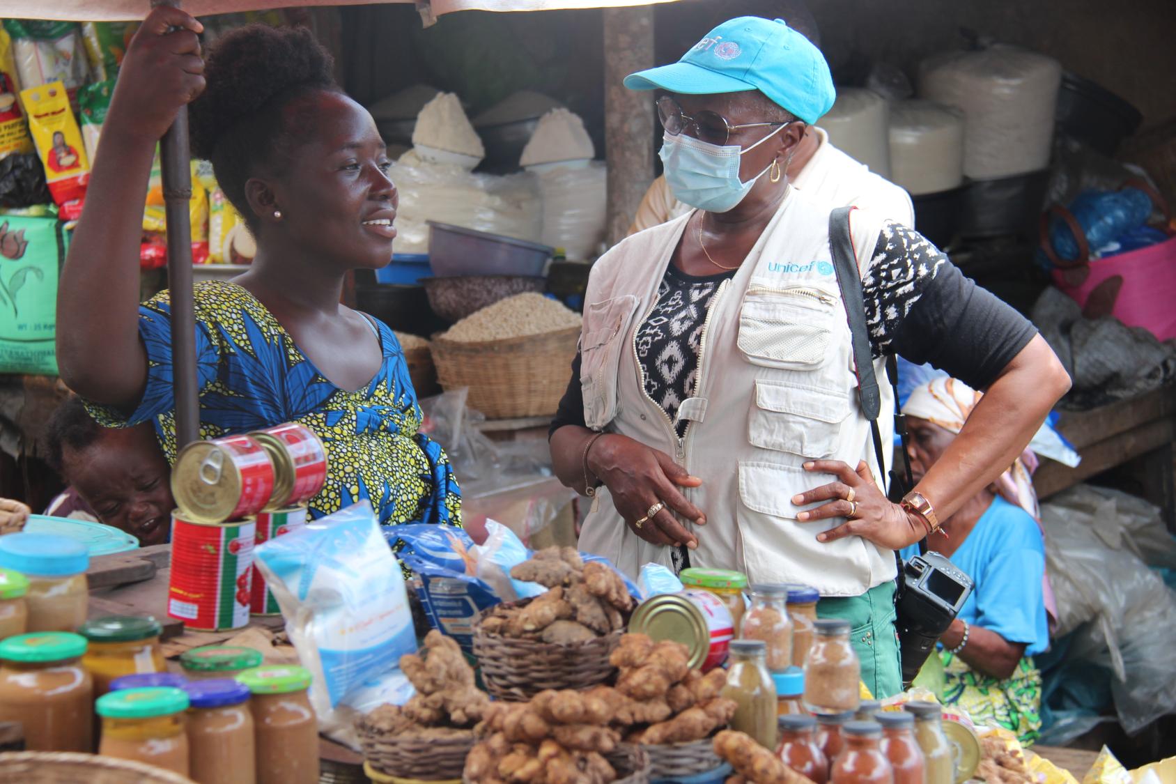 Une femme portant une casquette bleue, des lunettes de soleil et un masque chirurgical discute avec une autre femme devant un étal de maréché, au Bénin.
