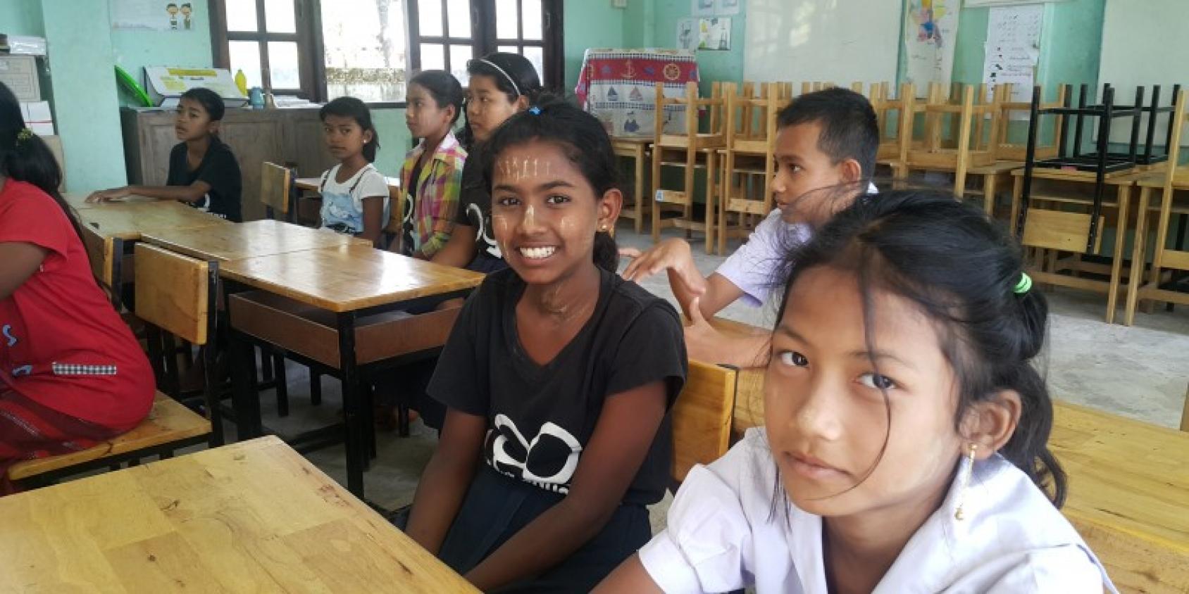 Девушки улыбаются в камеру, сидя в классной комнате. 