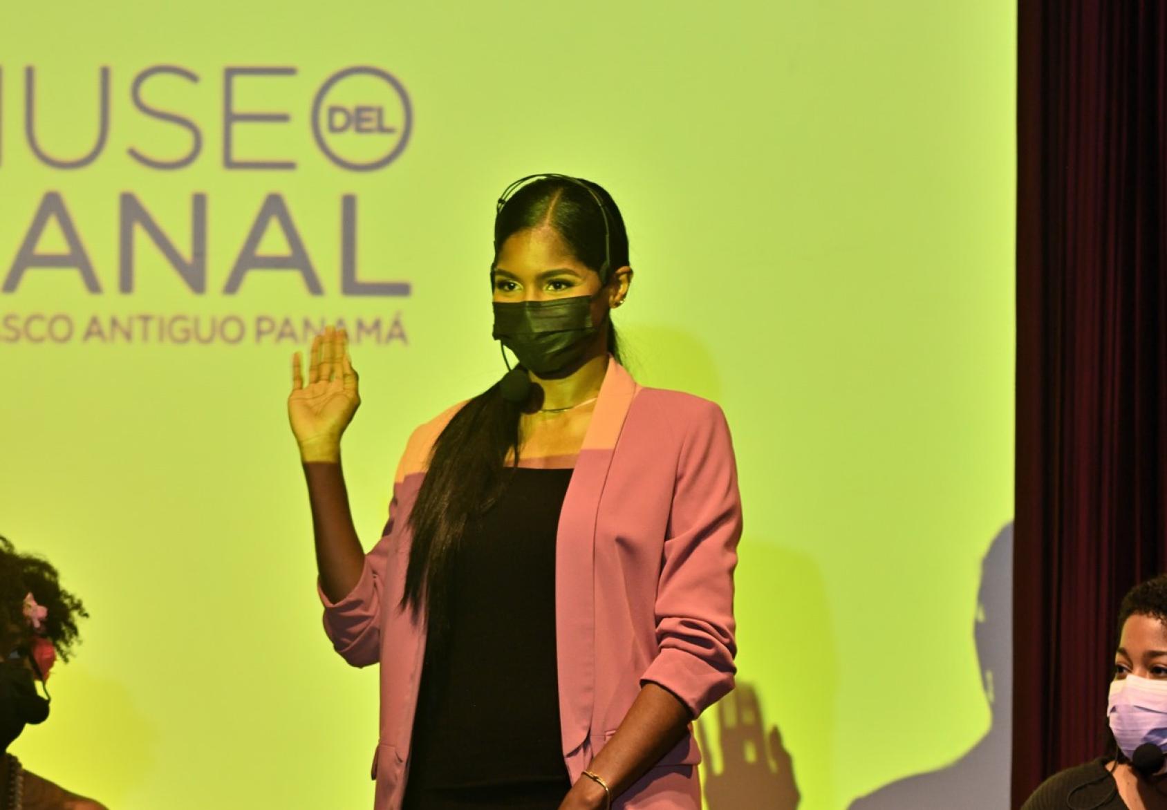 Una mujer con mascarilla negra levanta una mano a manera de saludo, sobre un escenario.
