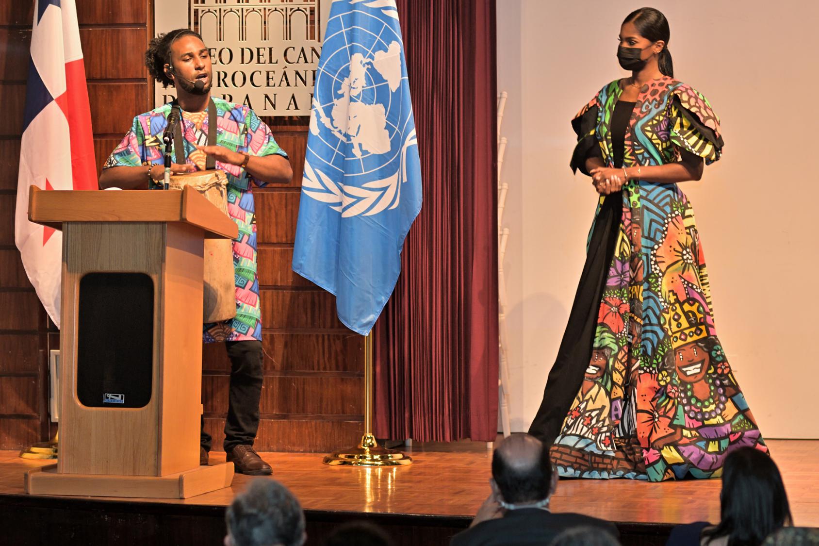 Un hombre y una mujer vestidos con ropas de colores están de pie en el escenario. El hombre canta y toca un tambor ante un micrófono.
