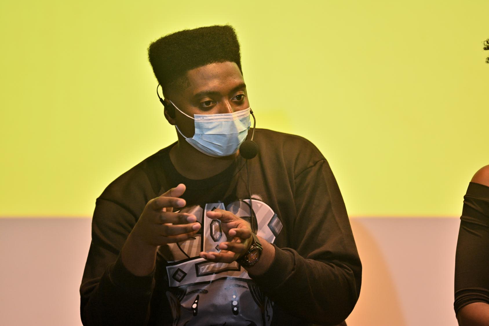 A man in a face mask speaks in front of a yellow projection screen. 