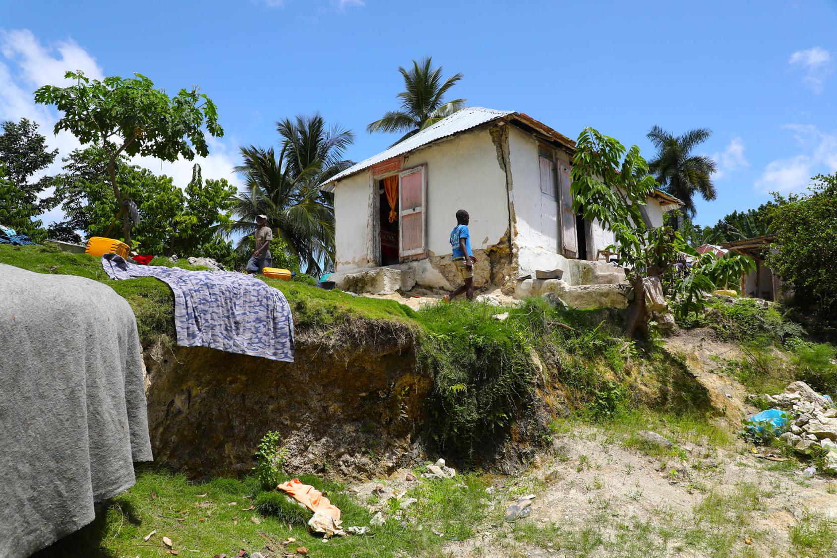 A person stands near a white home with several laundry items laid out to air dry on a hill. 