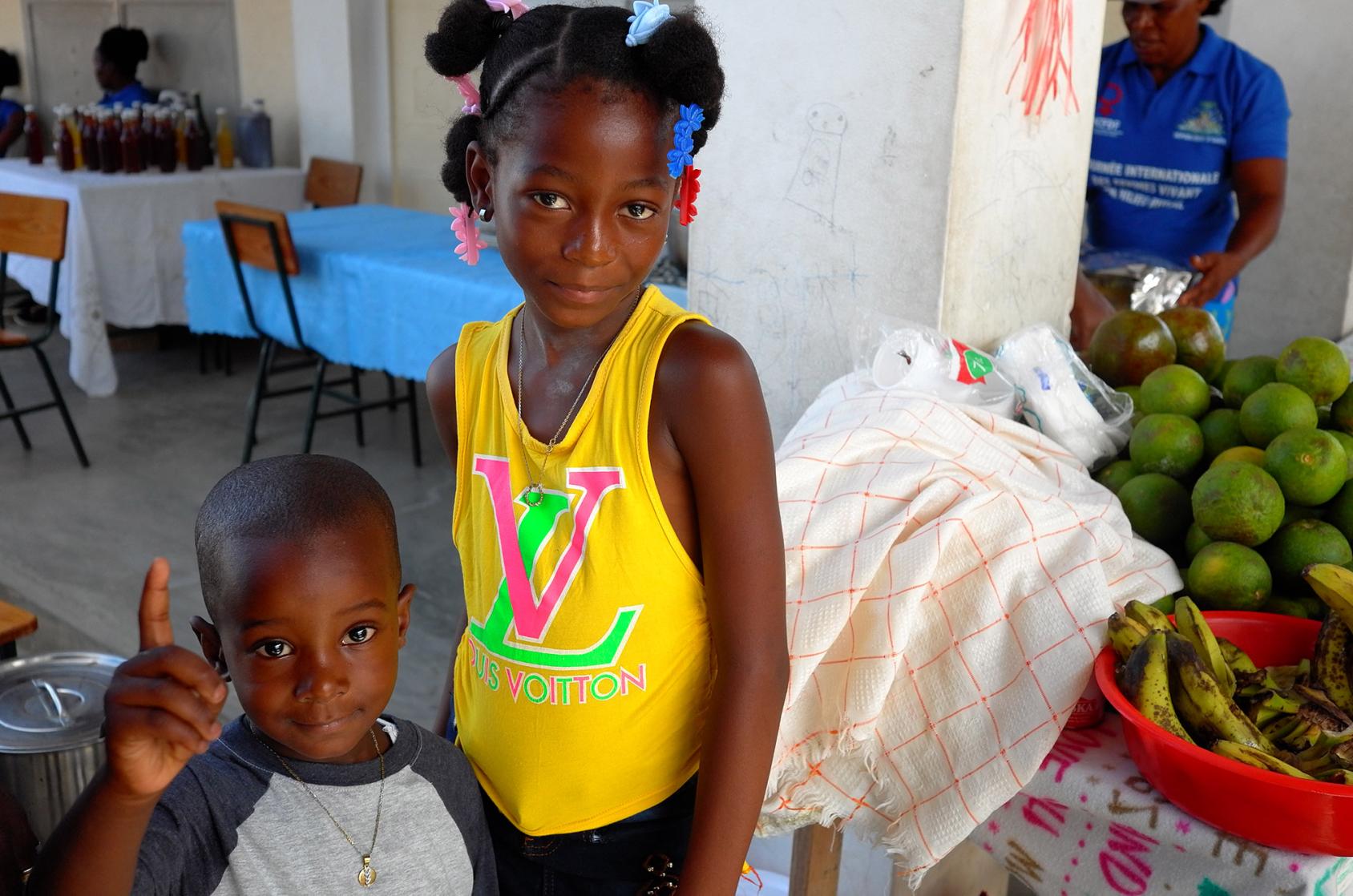 En Haïti, une fillette vêtue d’un débardeur jaune et un petit garçon se tiennnent debout à côté d'un stand de fruits en souriant à la caméra.