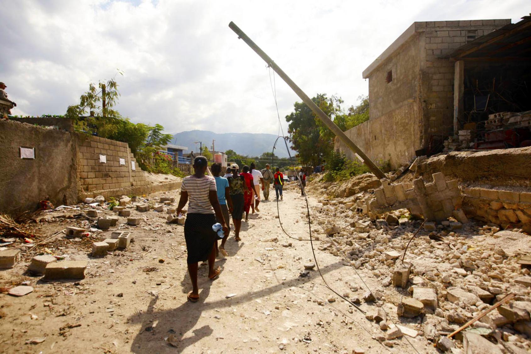 People walk in a line down a street with rubble on either side. 