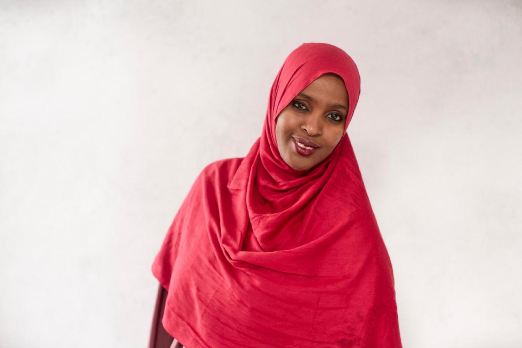 A smiling woman in a red hijab looks straight at the camera. 