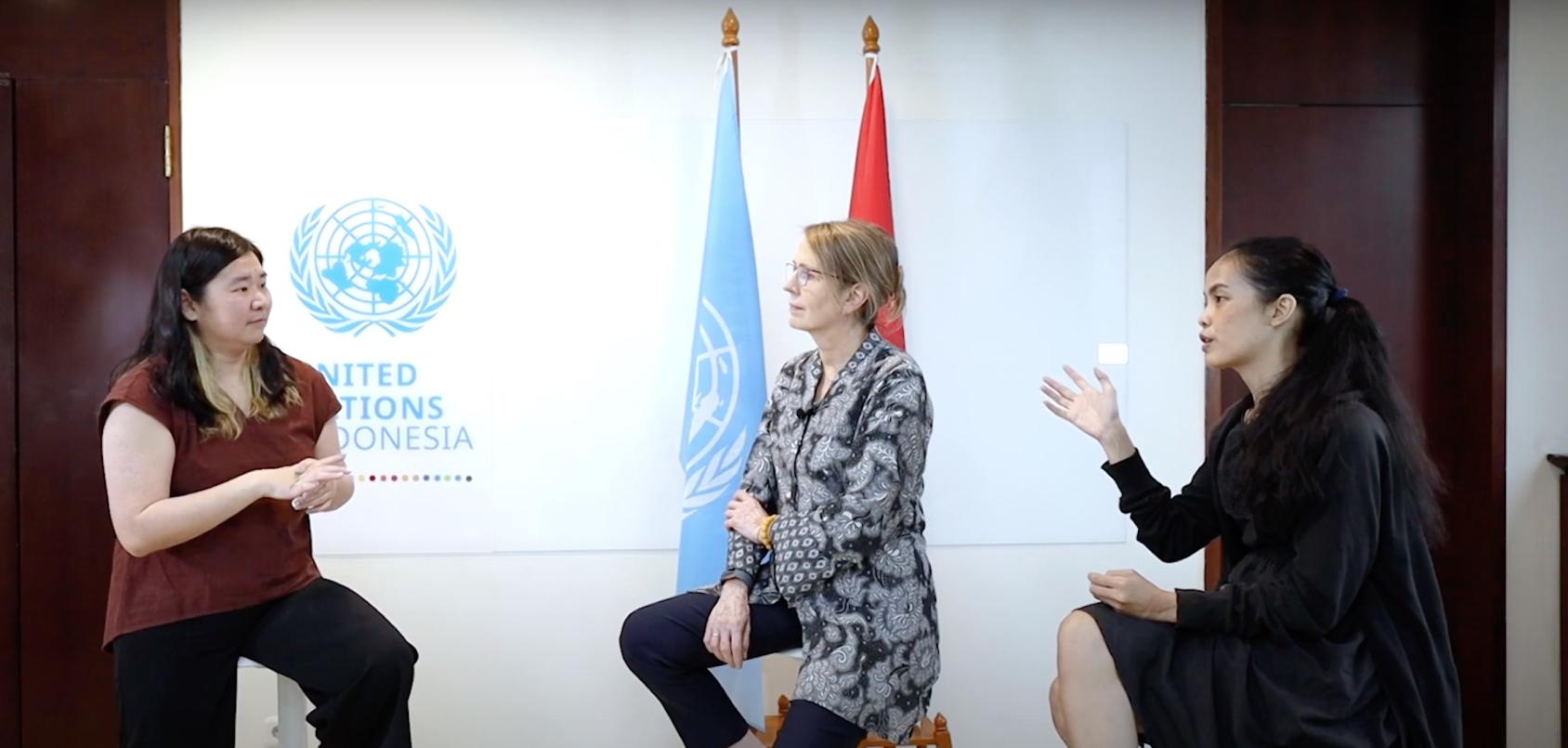 Tres mujeres mantienen una discusión frente a las banderas y el logotipo de las Naciones Unidas.
