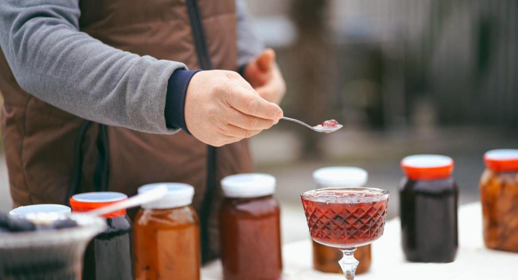 The close-up of a hand holding a spoon with jam with various jam jars in the background.