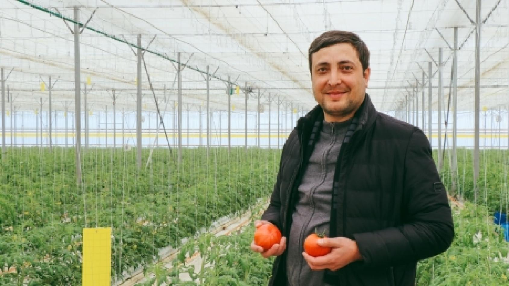 Un homme est photographié debout dans une serre, face caméra, tenant deux tomates dans les mains et souriant.