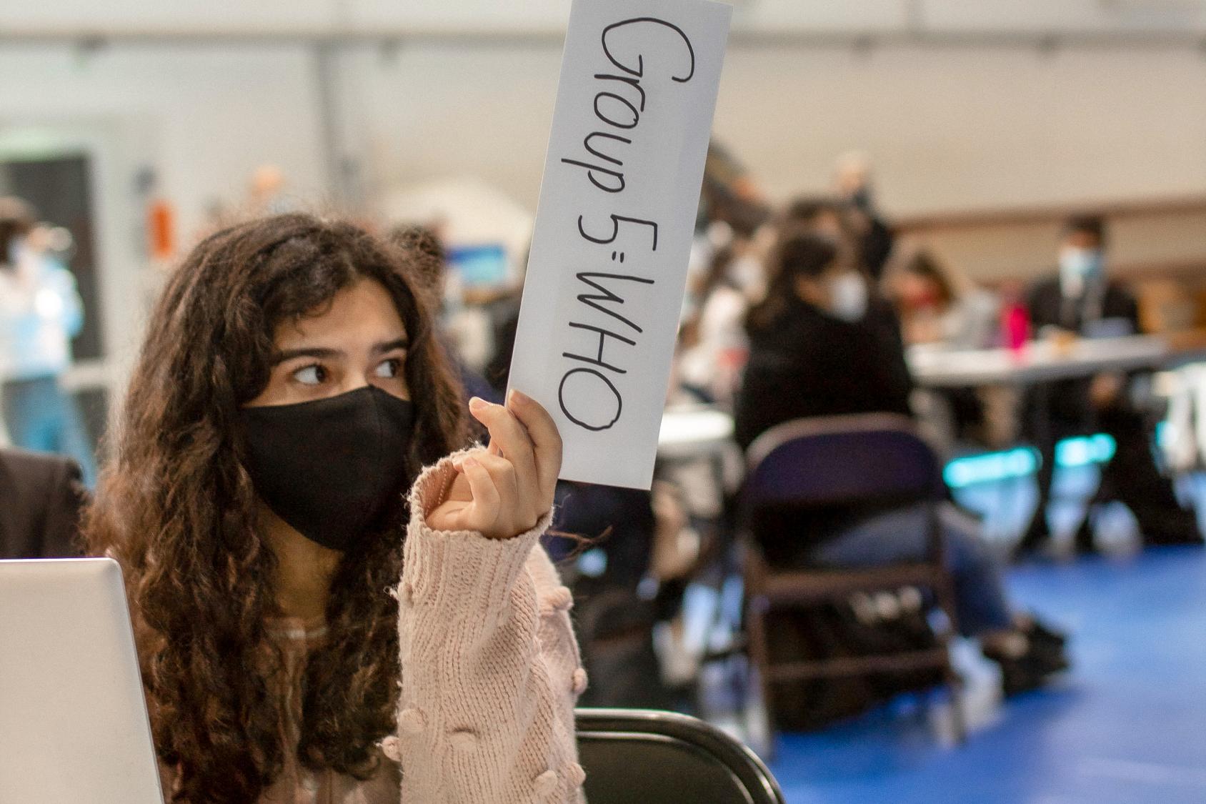 A girl in a black face mask holds a white sign up with one hand. 