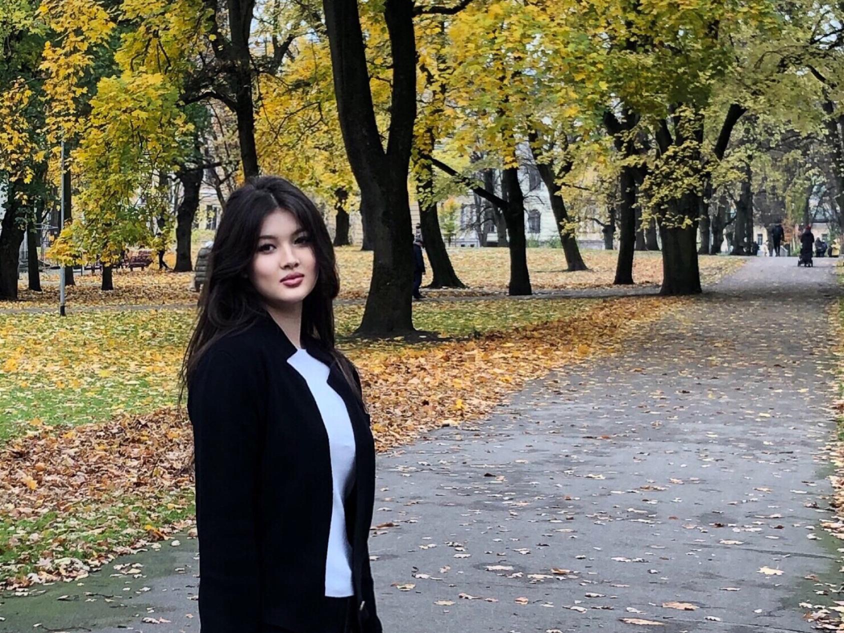 A girl in a black jacket stands outside in fall with fallen leaves behind her. 