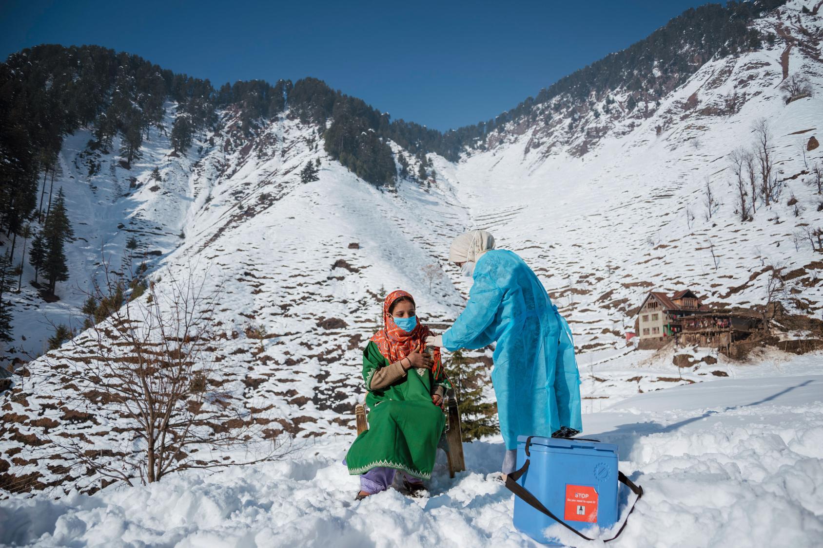 Une femme vêtue d’une robe verte se fait vacciner contre la COVID-19, près d’une glacière, par une agente de santé vêtue d’une blouse de protection bleue, au sommet d'une montagne enneigée, en Inde.