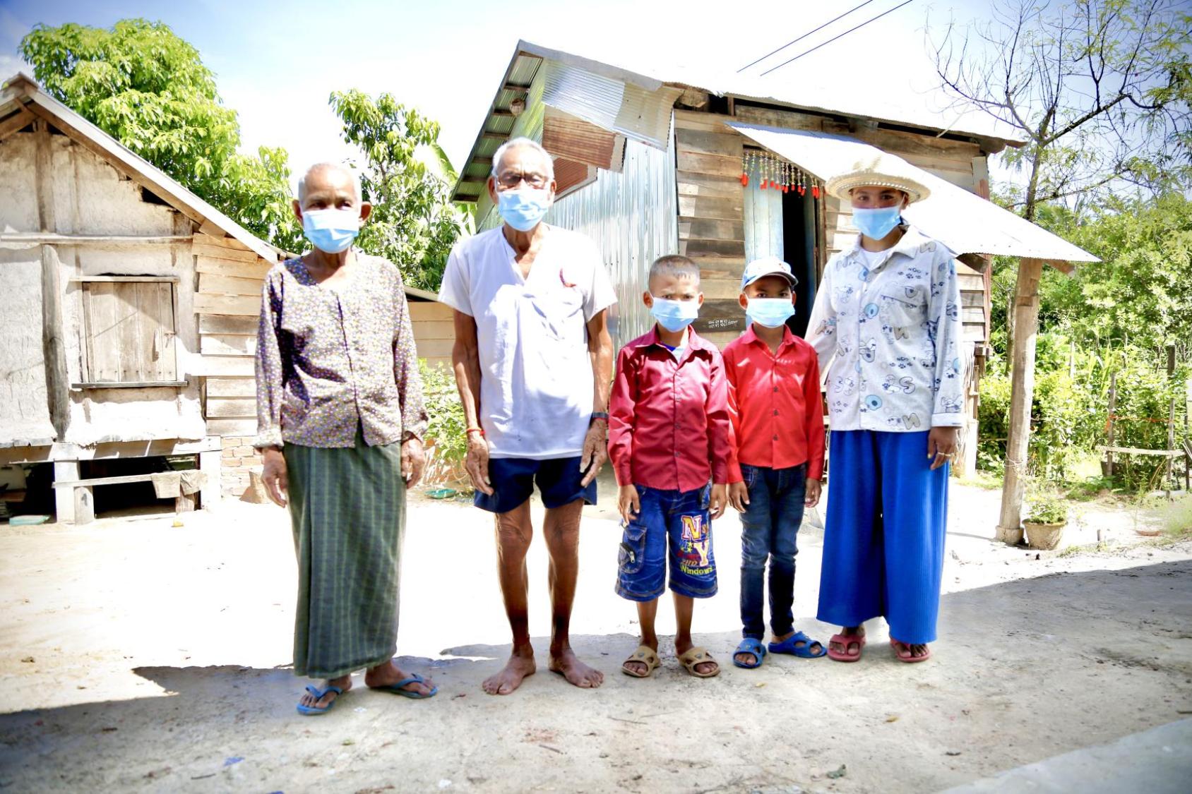 Les membres d’une famille cambodgienne portant chacun un masque de protection respiratoire se tiennent de debout, côte à côte, face caméra, devant de petites maisons en bois, par une journée ensoleillée.