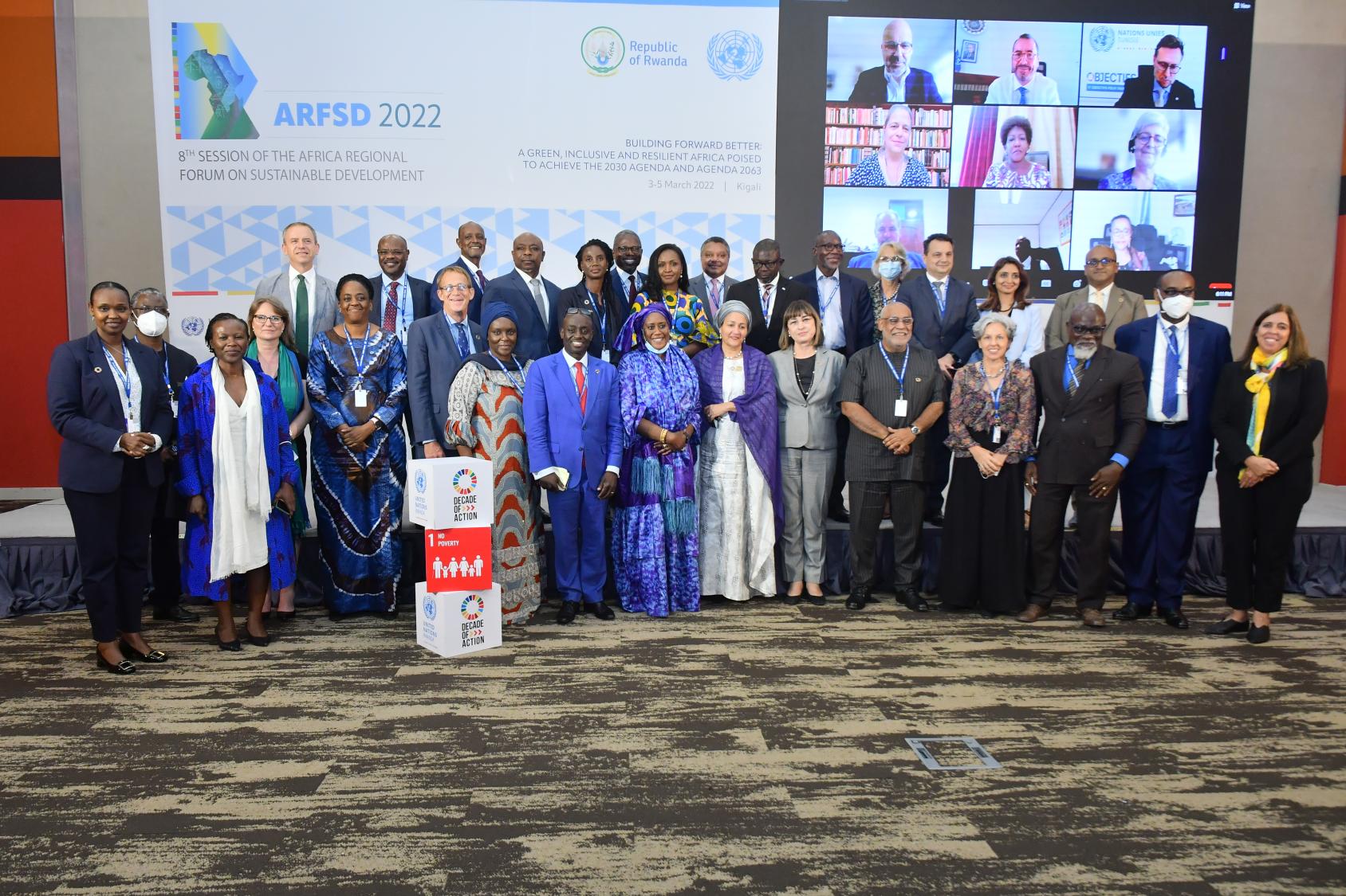 Foto grupal de la Vicesecretaria General junto a los 29 Coordinadores Residentes presentes físicamente en el Centro de Convenciones de Kigali, con una pantalla atrás donde se ve a más asistentes conectados de forma virtual.