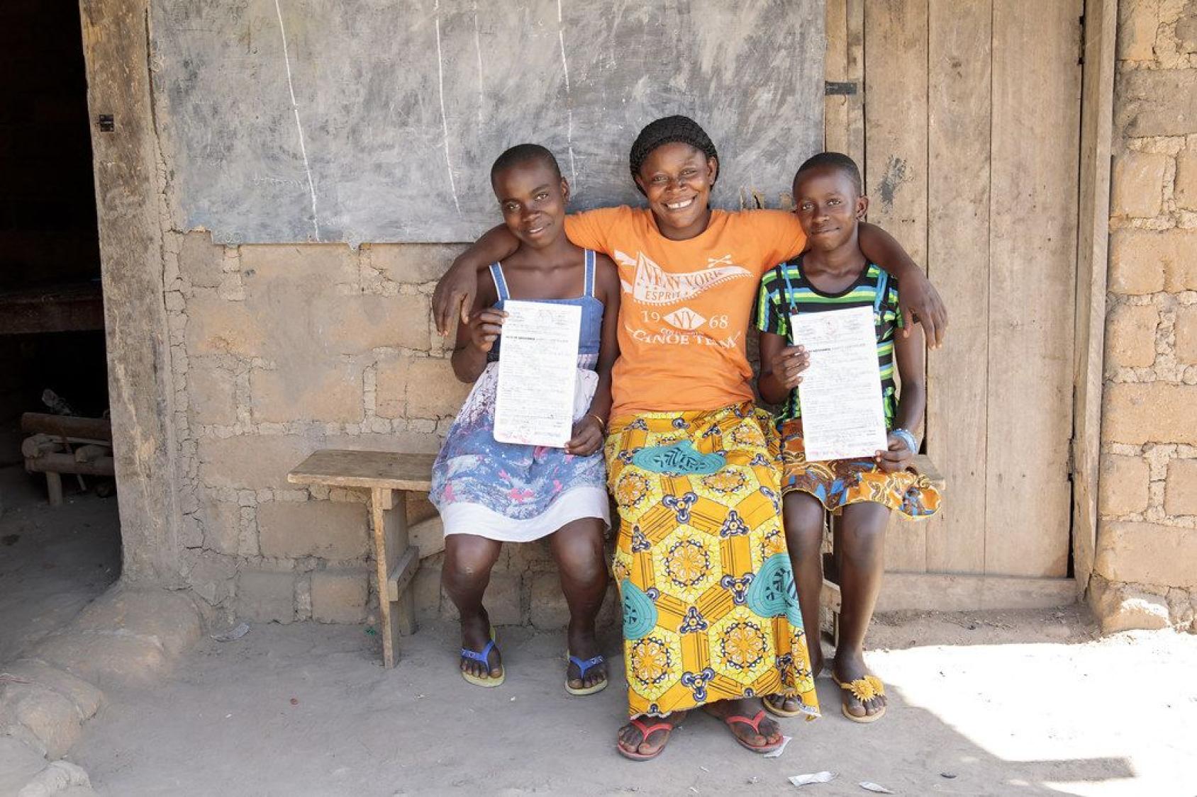 Two girls from Cameroon sit with their mother on a bench outside a house, proudly showing off their newly issued birth certificates, and smiling at the camera. 