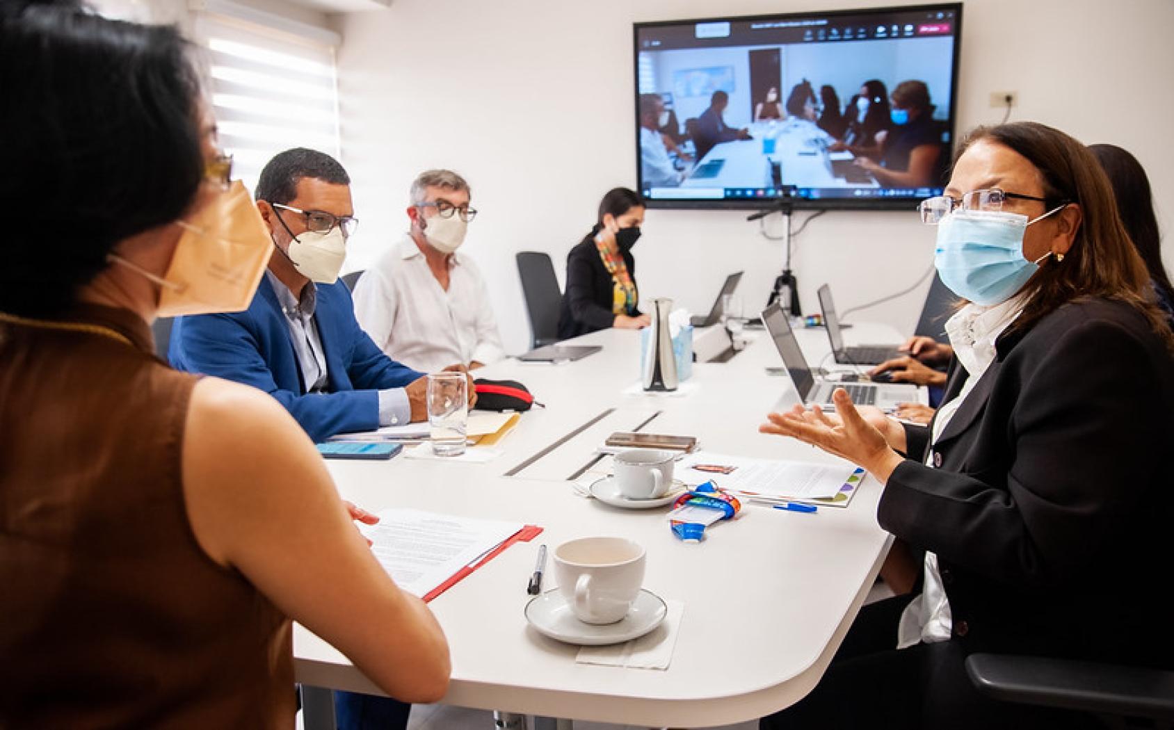 Miembros del personal de Naciones Unidas en una reunión de trabajo con una videoconferencia en el fondo.